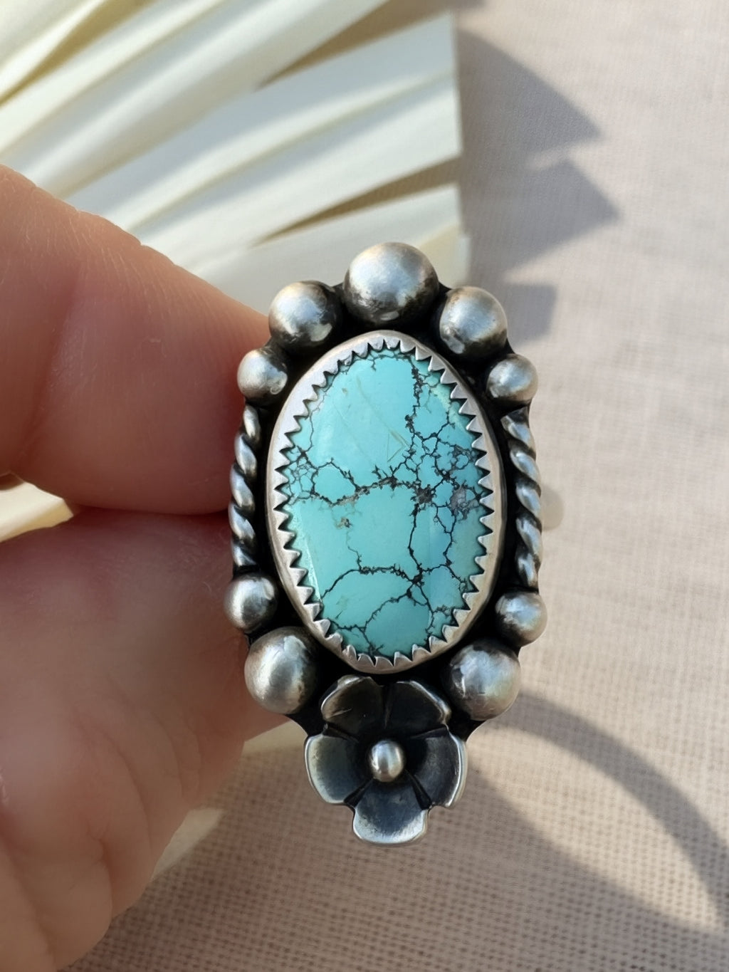 A hand holding a sterling silver ring featuring an oval turquoise cabochon with prominent black matrix veining, surrounded by a beaded border and a flower motif at the bottom.