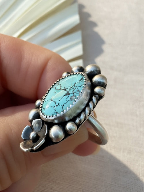 A hand holding a silver ring with a large oval turquoise stone and decorative silver beads around the setting, photographed against a light neutral background.