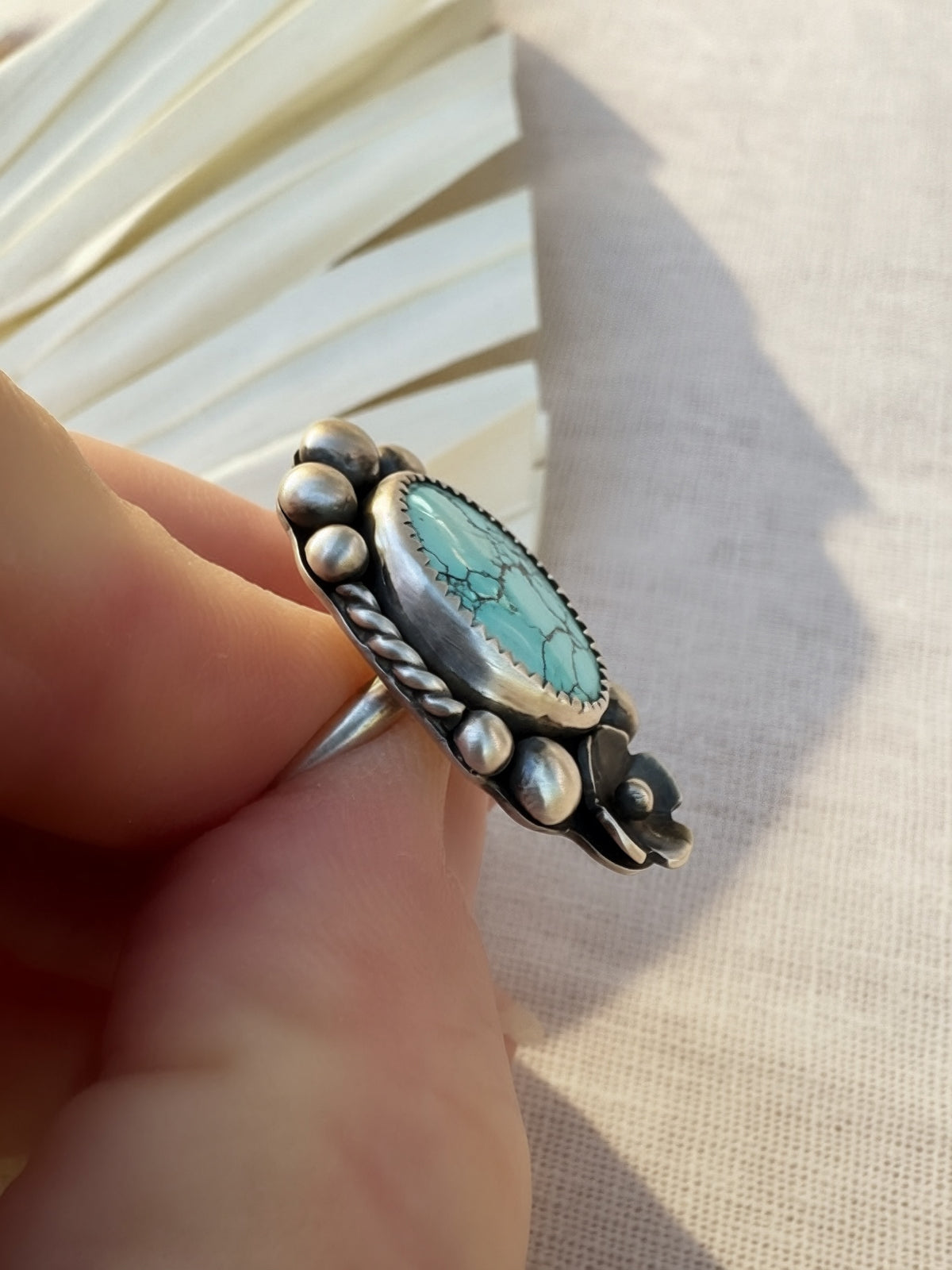 A hand holding a silver ring featuring an oval turquoise stone with natural matrix patterns, set in a decorative bezel with small silver beads.