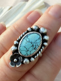 A sterling silver ring with an oval turquoise stone and a floral accent, shown worn on a person's finger.