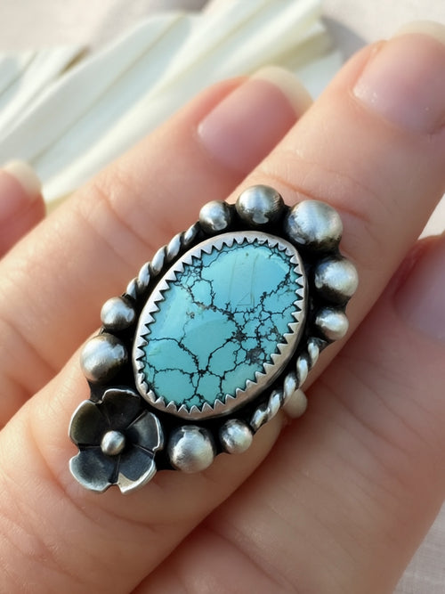 A sterling silver ring with an oval turquoise stone and a floral accent, shown worn on a person's finger.
