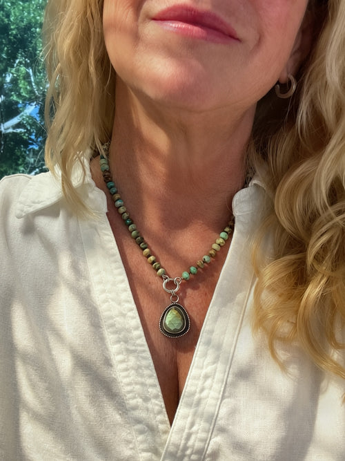 A close-up of a woman wearing a turquoise beaded necklace with a green and gold labradorite teardrop pendant, paired with a white collared shirt.