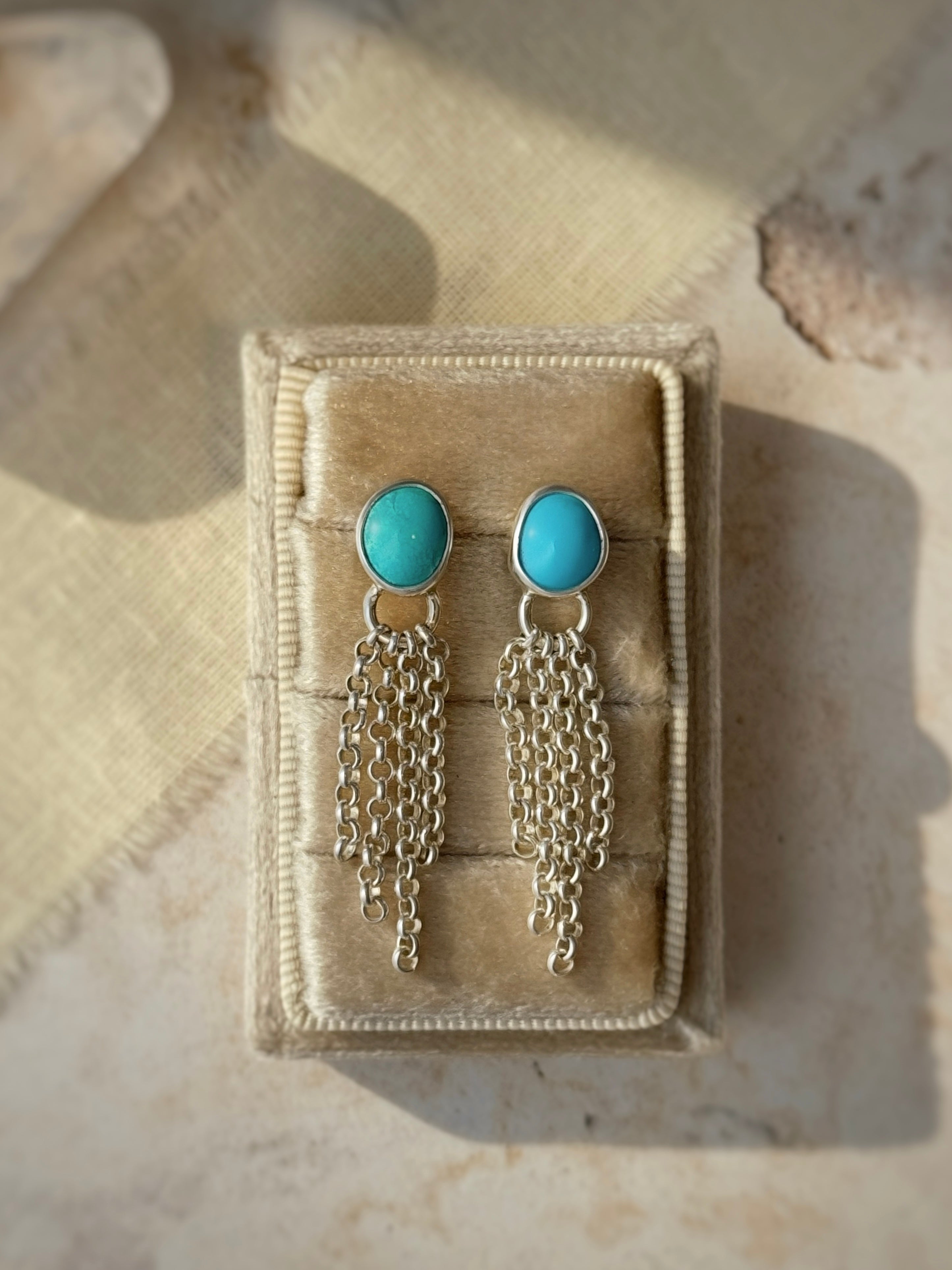 Turquoise earrings with silver chains on a beige jewelry box.