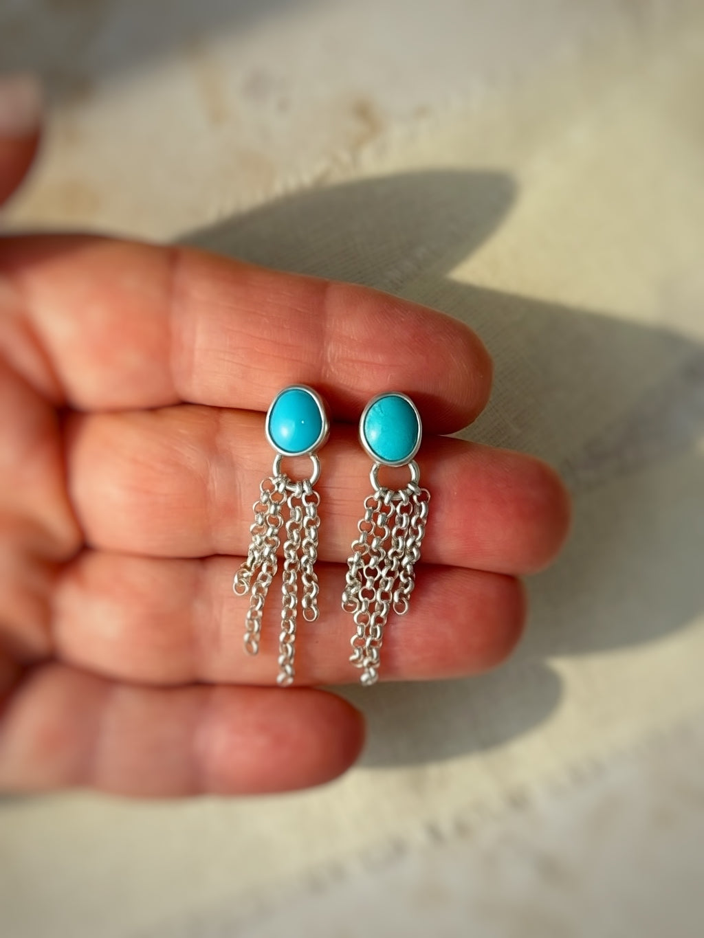 Turquoise stone earrings with silver chains held in a hand against a neutral background