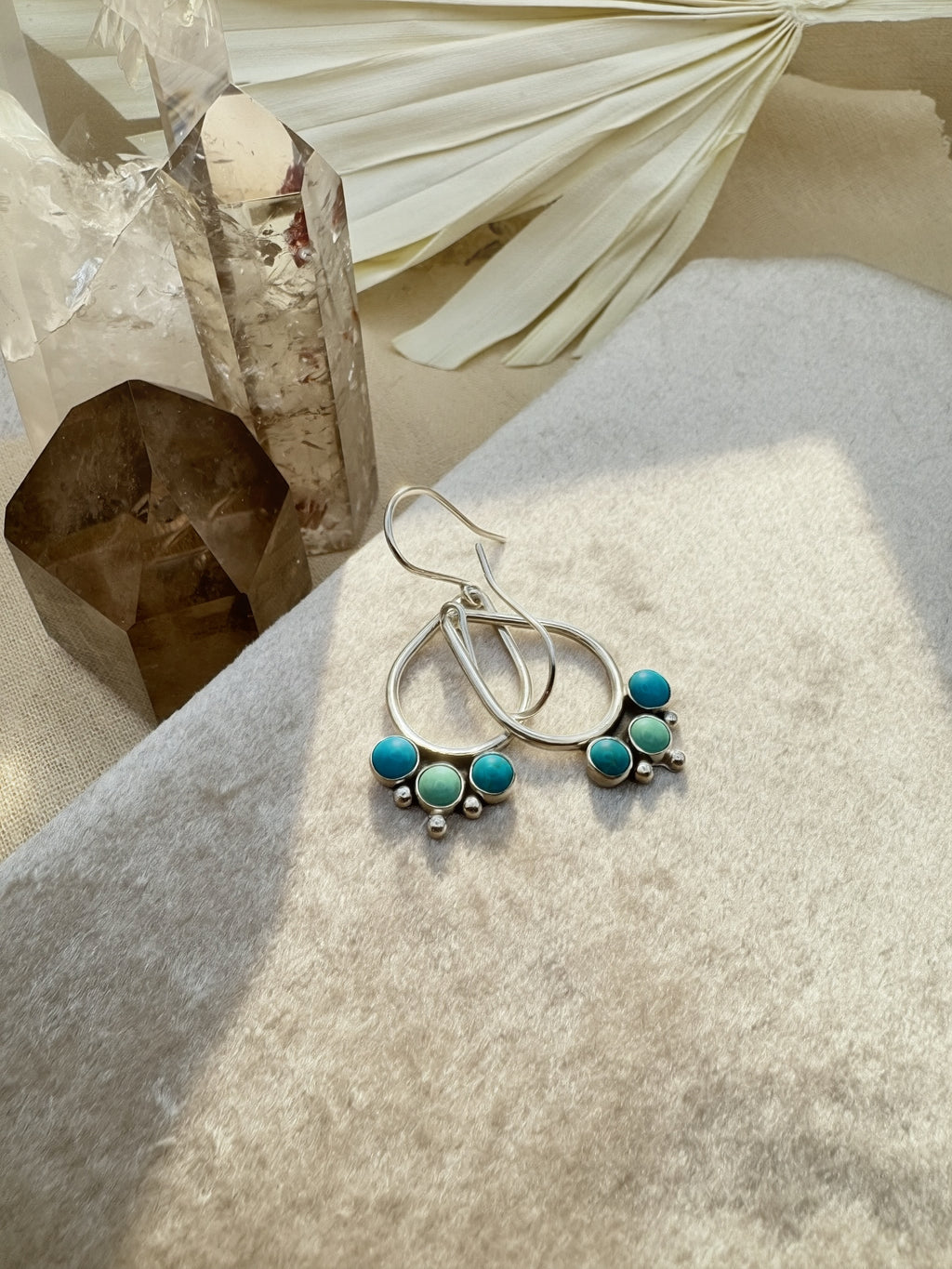 Hand-fabricated sterling silver open teardrop earrings with three bezel-set Kingman turquoise cabochons and silver granule detail, on a neutral background with crystal and dried palm