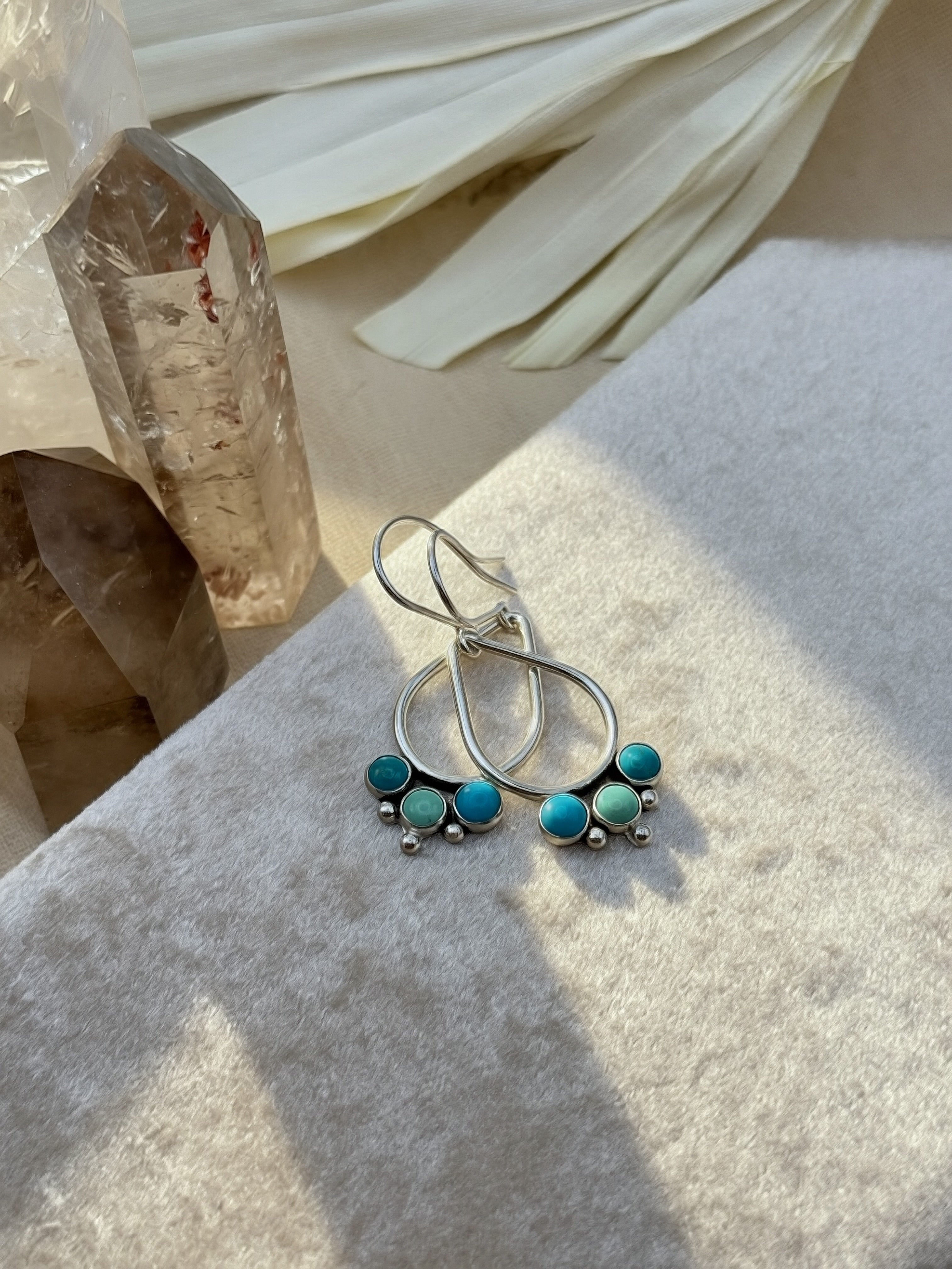 Hand-fabricated sterling silver open teardrop earrings with three bezel-set Kingman turquoise cabochons and silver granule detail, on a neutral background with crystal and dried palm