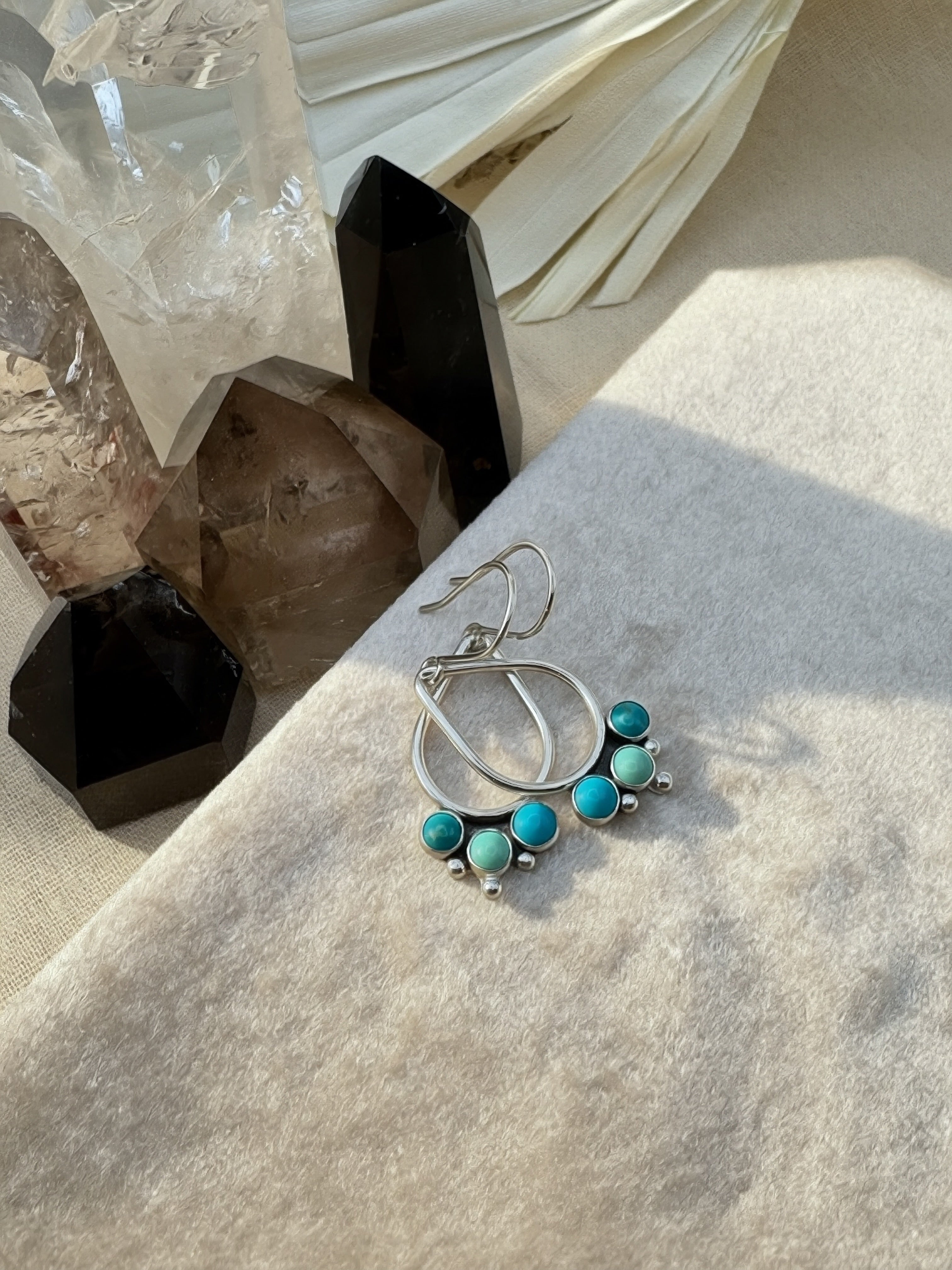 Hand-fabricated sterling silver open teardrop earrings with three bezel-set Kingman turquoise cabochons and silver granule detail, on a neutral background with crystal and dried palm