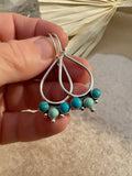 Hand-fabricated sterling silver open teardrop earrings with three bezel-set Kingman turquoise cabochons and silver granule detail, held against crystal and dried palm