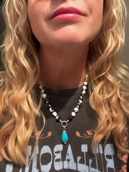A close-up of a woman wearing a beaded necklace with a turquoise pendant. The necklace features white and black Buffalo beads with a silver charm keeper ring and a teardrop-shaped AZ turquoise pendant.