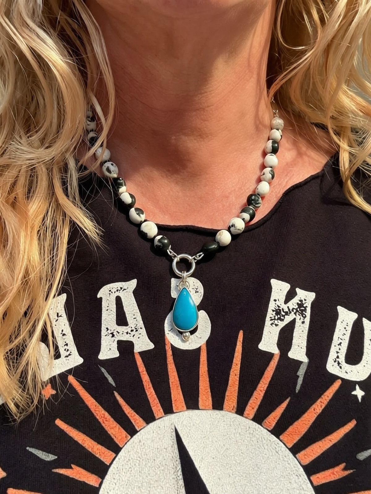 A close-up of a woman wearing a beaded necklace with a turquoise pendant. The necklace features white and black white Buffalo beads with a silver charm keeper ring and a teardrop-shaped AZ turquoise pendant.