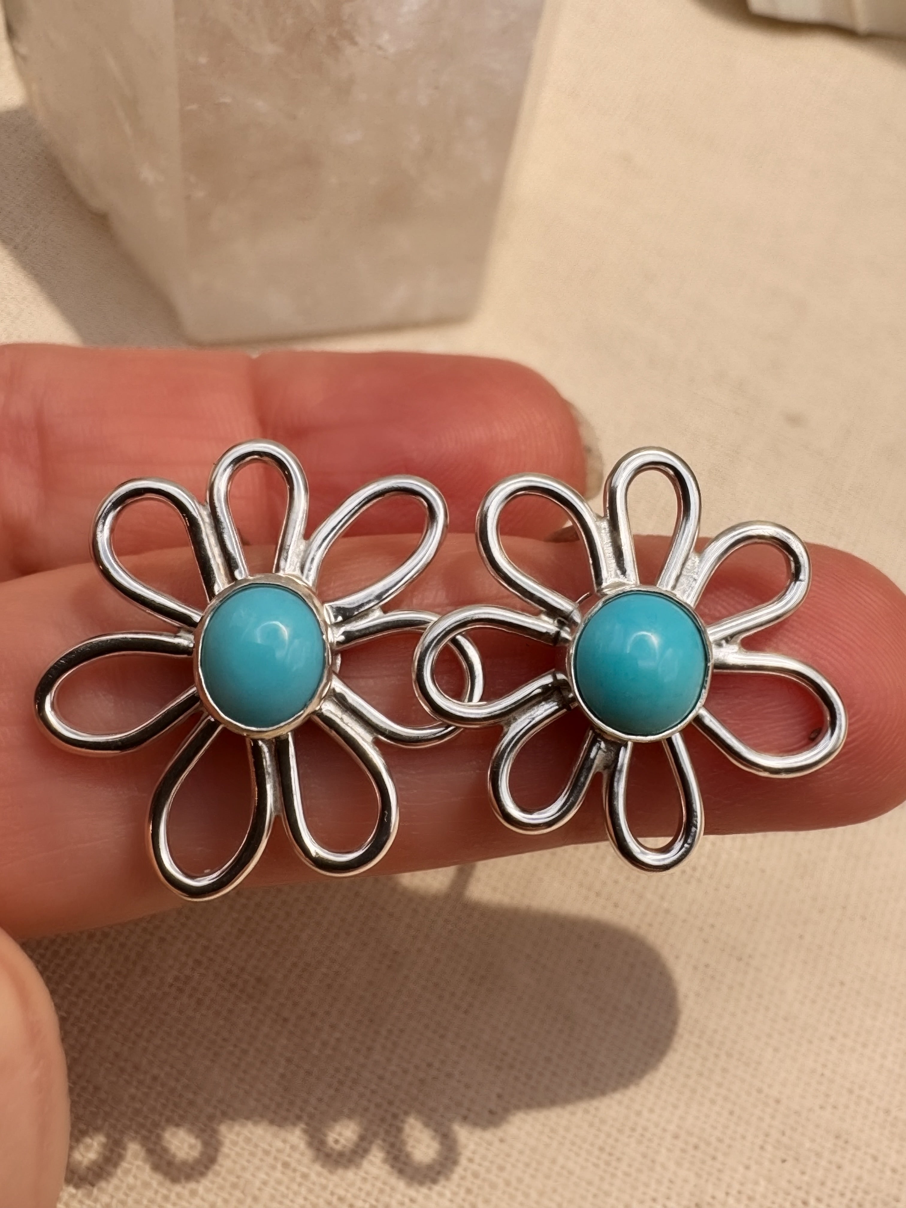 Silver flower-shaped earrings with turquoise centers held between fingers on a beige background