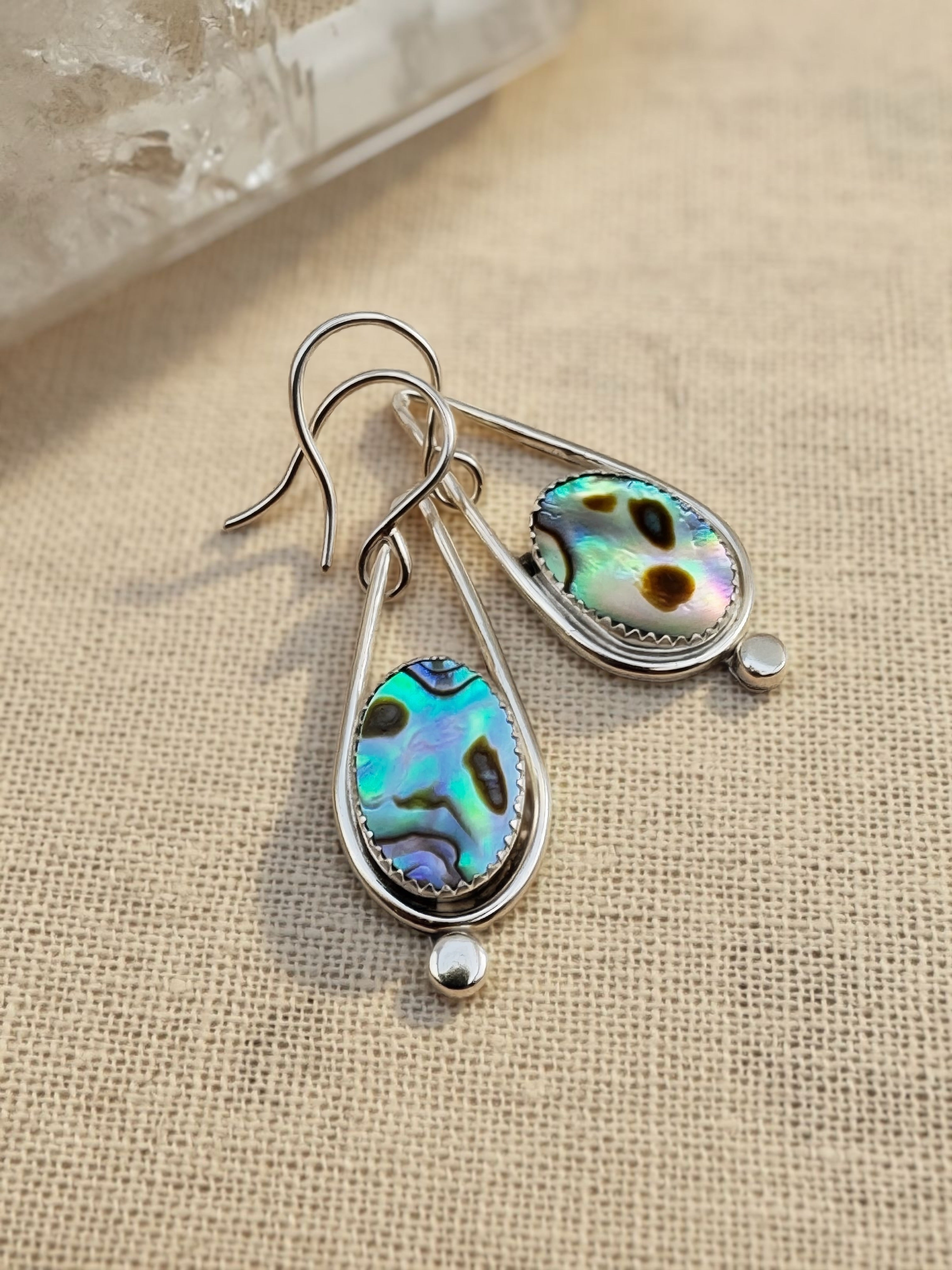 Pair of abalone shell teardrop earrings in sterling silver with French hook ear wires, displayed on natural linen