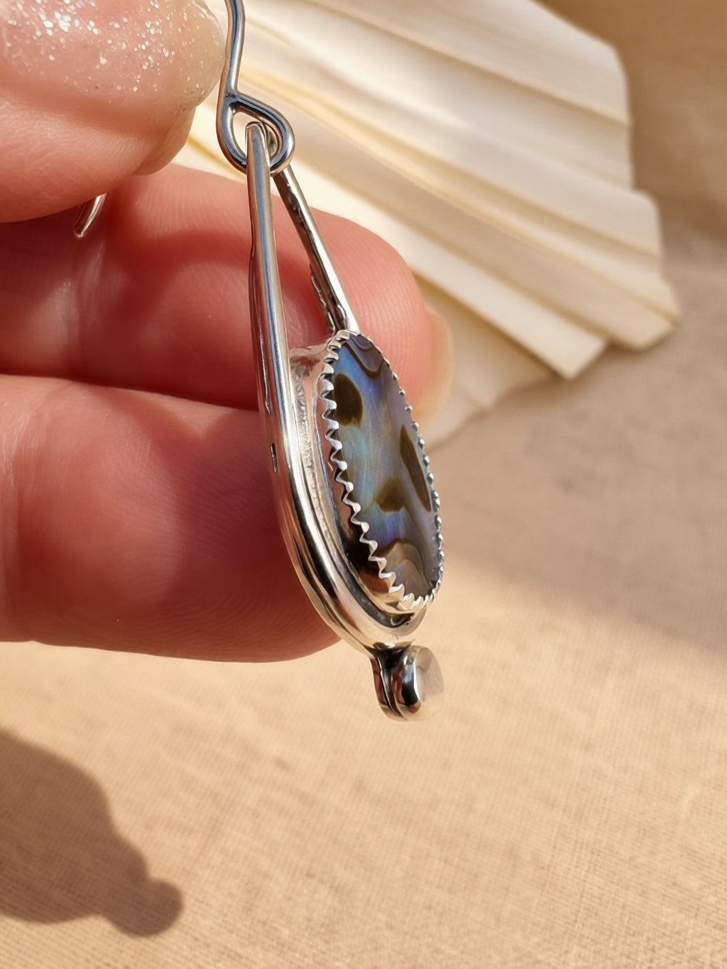 Side view Close-up of handmade abalone shell drop earring held in hand, showing iridescent blue and green natural shell in sterling silver bezel