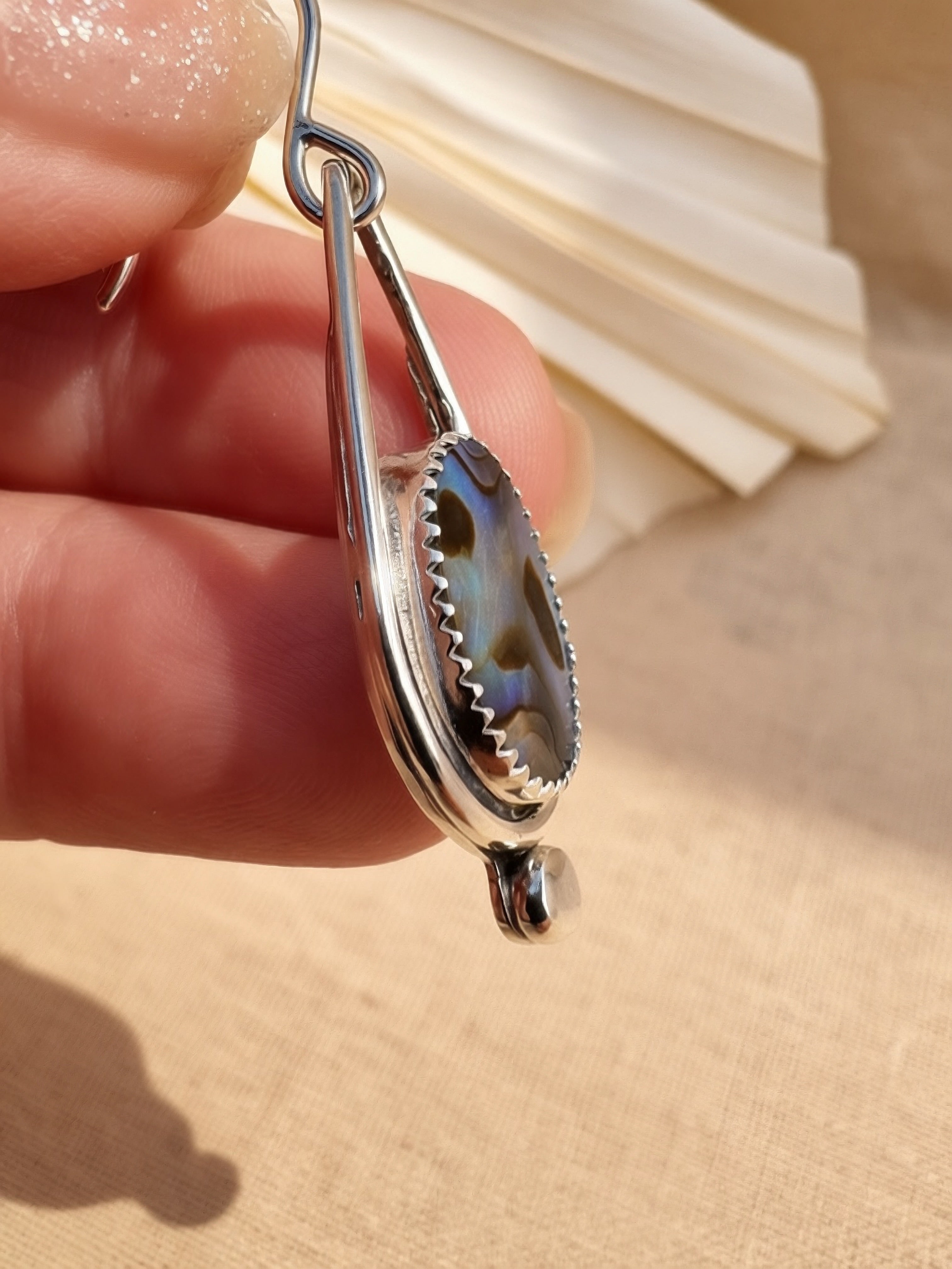 Side view Close-up of handmade abalone shell drop earring held in hand, showing iridescent blue and green natural shell in sterling silver bezel