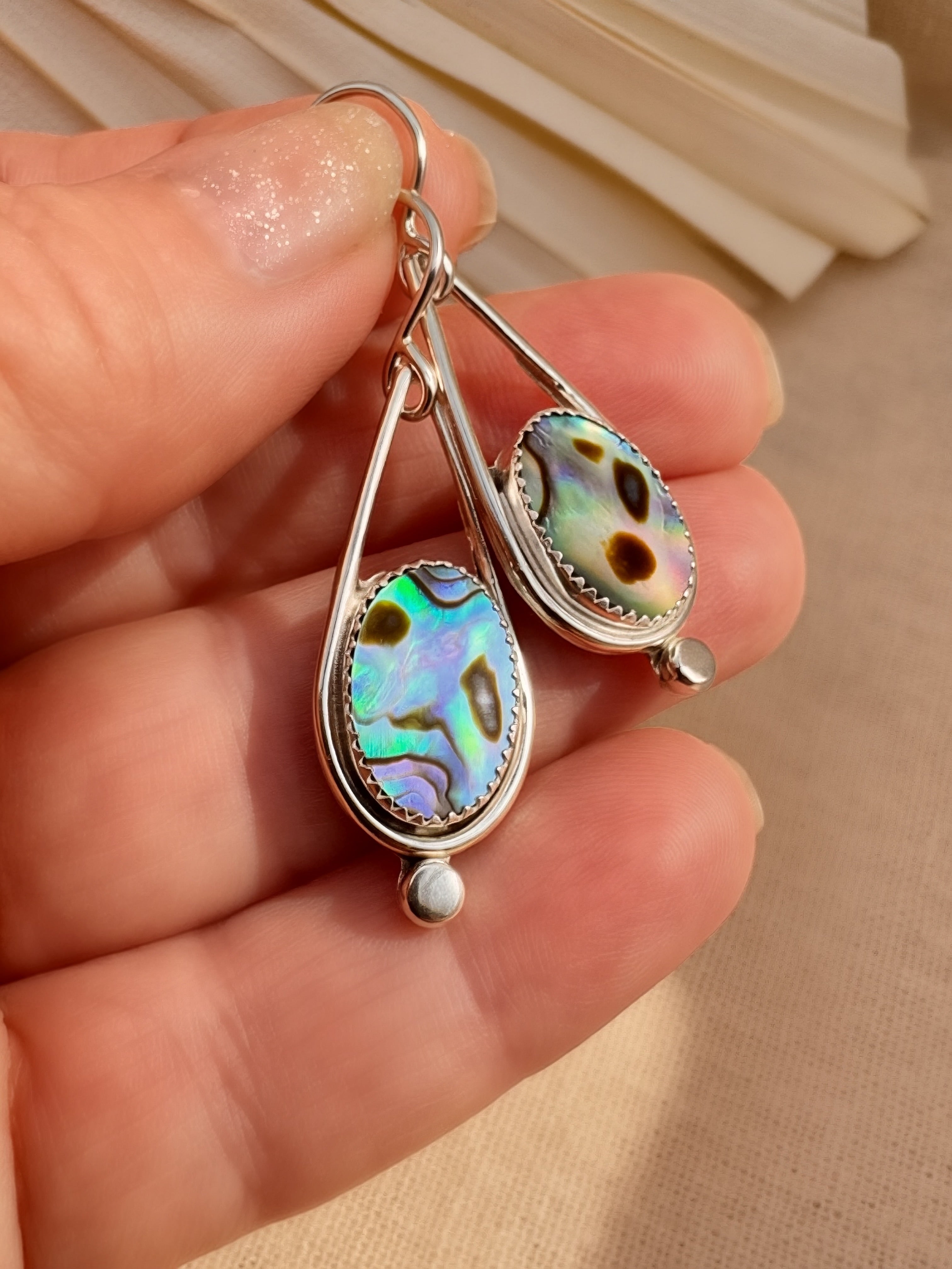 Close-up of handmade abalone shell drop earring held in hand, showing iridescent blue and green natural shell in sterling silver bezel