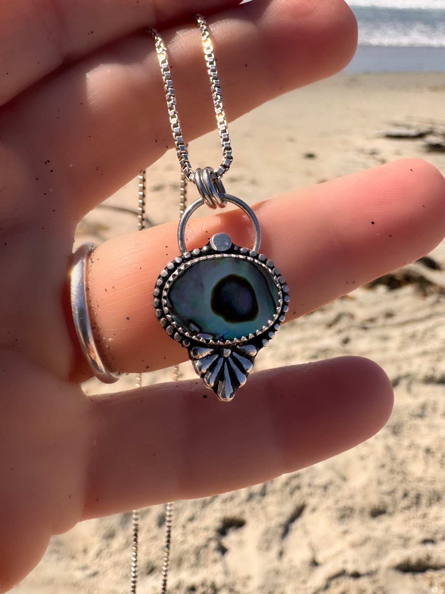 Silver and abalone necklace with an eye-shaped pendant held by a hand on a beach.