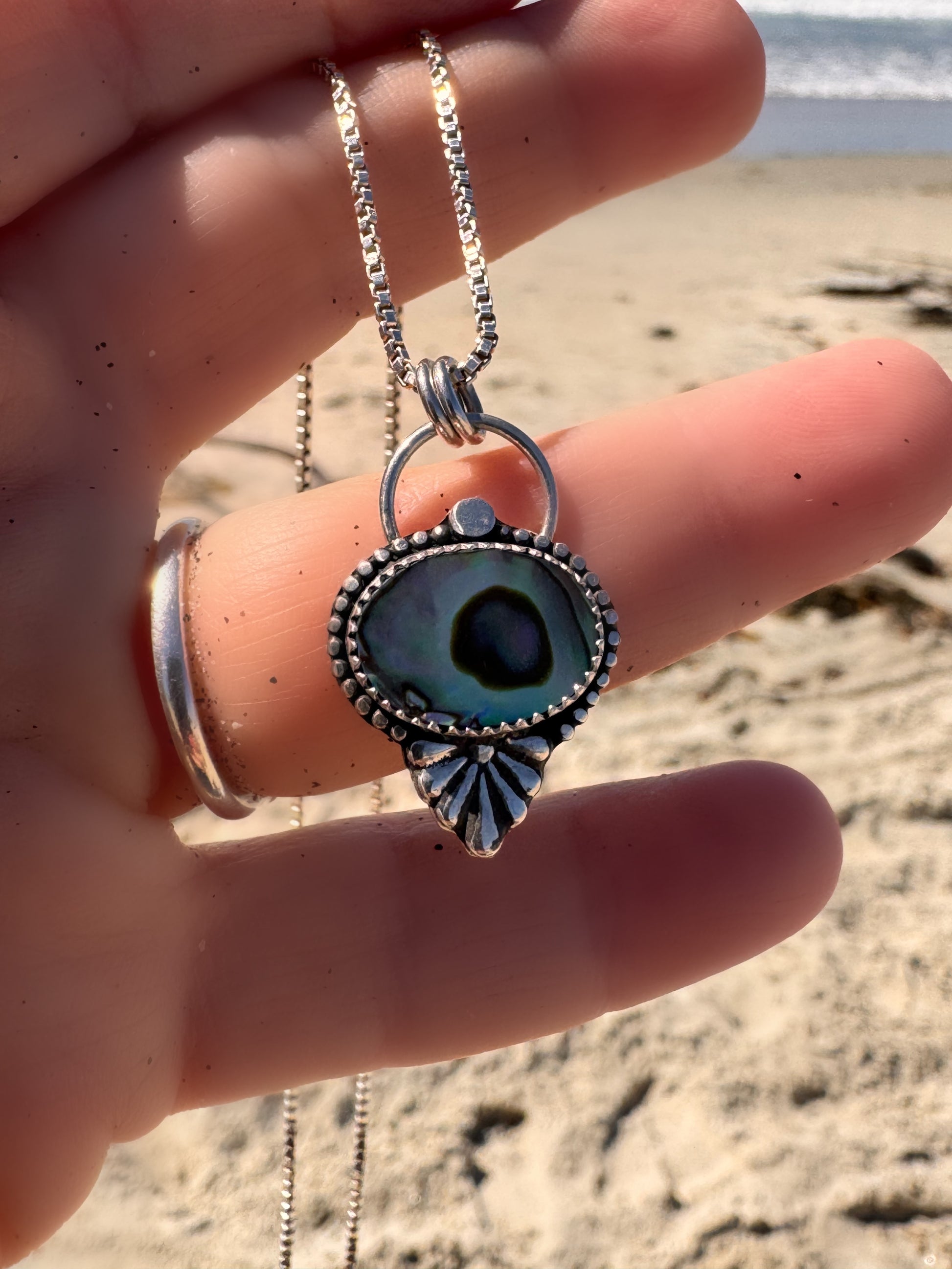 Silver and abalone necklace with an eye-shaped pendant held by a hand on a beach.