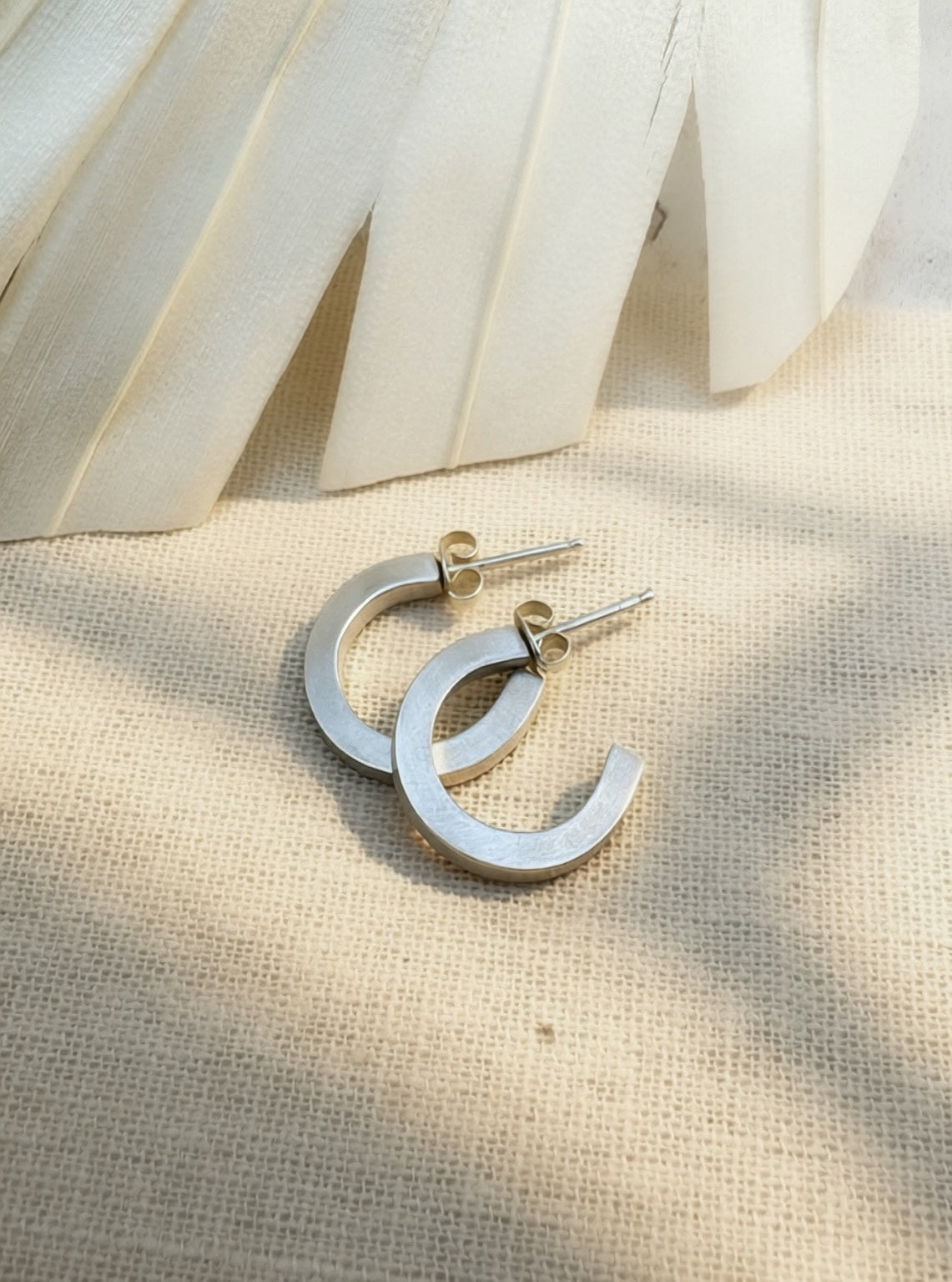 Satin sterling silver hoop earrings on cream fabric, showing flat faces, squared edges, posts, and butterfly backs.