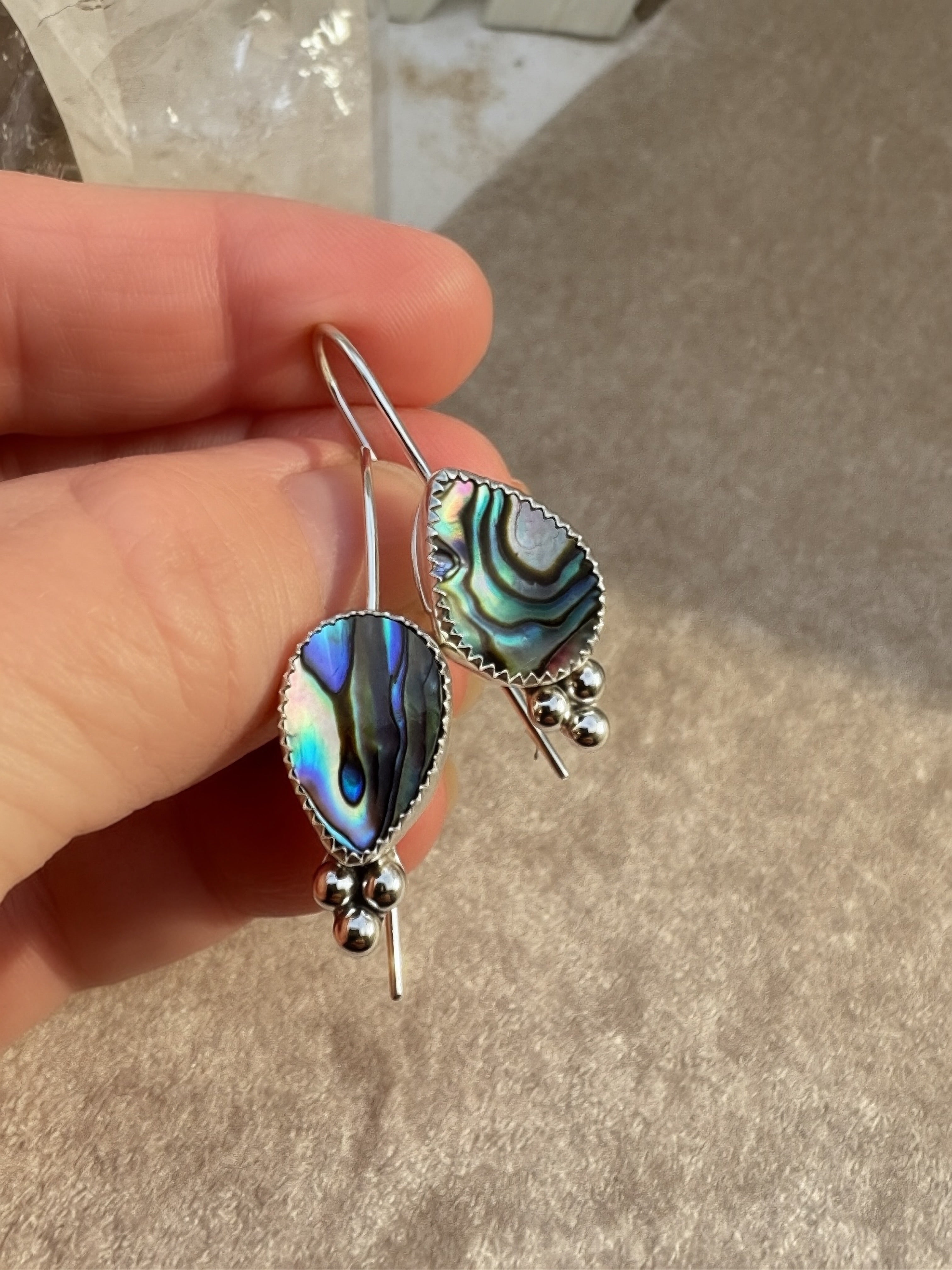 A close-up of a hand holding a pair of silver dangle earrings with iridescent abalone shell inlays and small silver bead drops, against a neutral textured background.