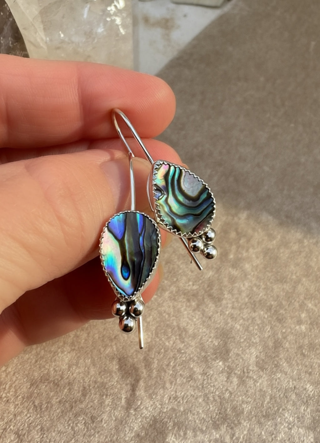 A hand holding a pair of silver dangle earrings with teardrop-shaped abalone shell pendants showing iridescent blue, green, and black patterns, with small silver bead clusters below.
