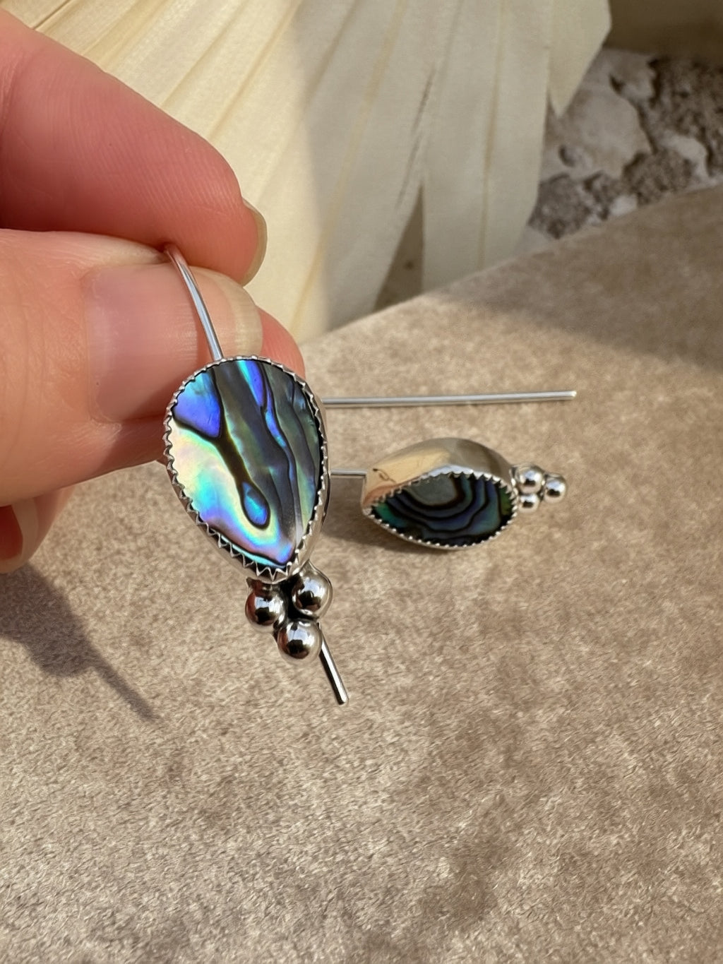 A hand holding a pair of silver dangle earrings with teardrop-shaped abalone shell inlays. The earrings feature a silver bezel setting, a vibrant iridescent abalone center, and small silver beads at the bottom, shown against a neutral textured background.