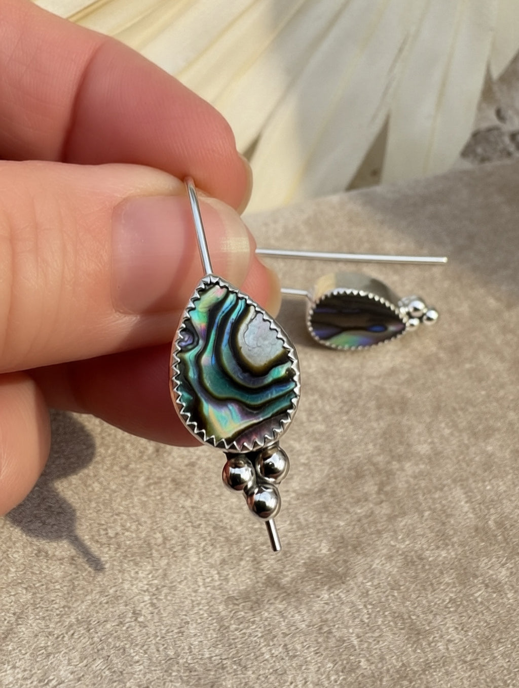 A close-up of a hand holding a silver teardrop earring with an iridescent abalone shell inlay, with a matching earring resting on a neutral surface in the background.