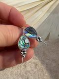A close-up of a hand holding a pair of silver dangle earrings with teardrop-shaped abalone shell inlays that show iridescent green, blue, and purple hues, set against a neutral draped fabric background.