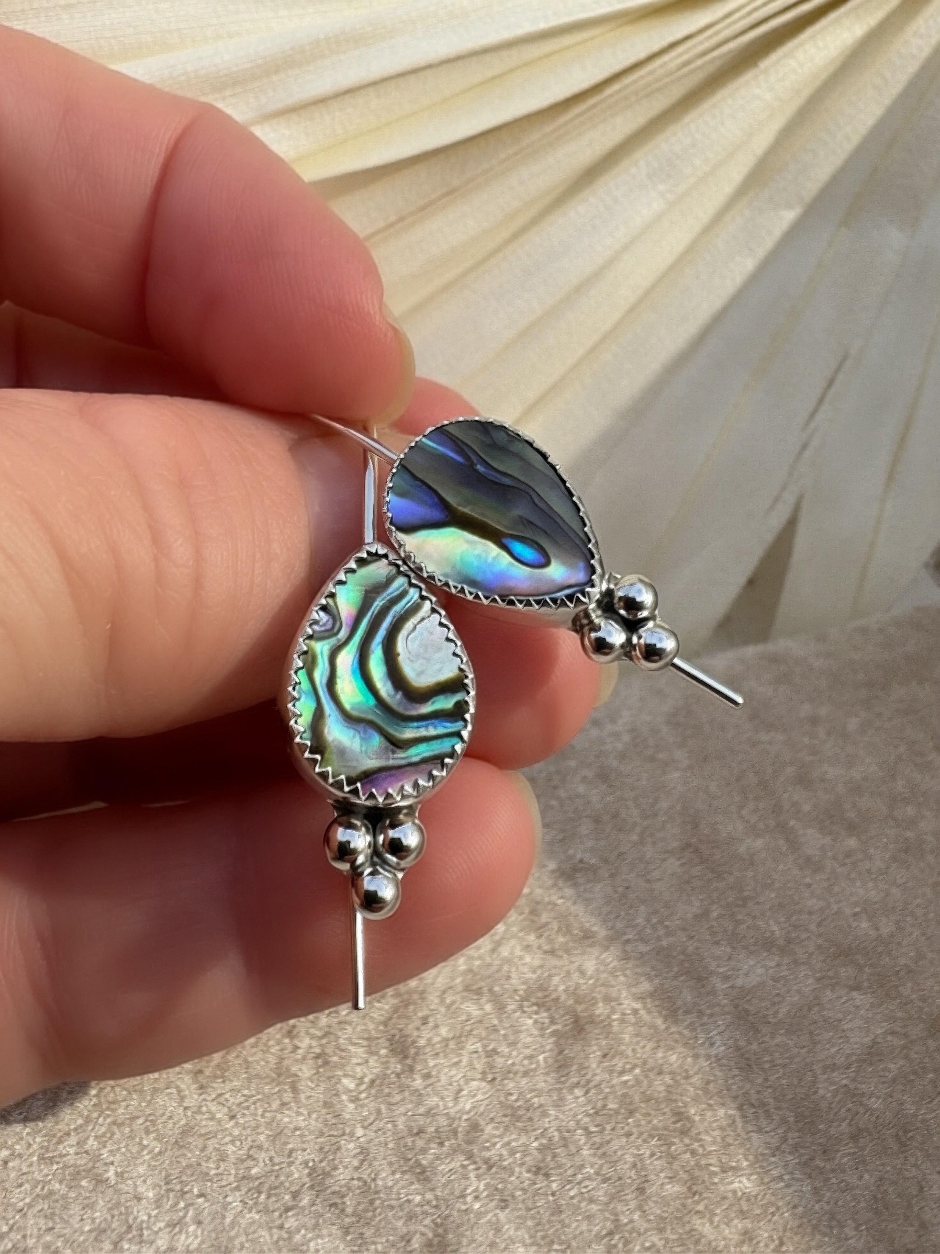 A close-up of a hand holding a pair of silver dangle earrings with teardrop-shaped abalone shell inlays that show iridescent green, blue, and purple hues, set against a neutral draped fabric background.