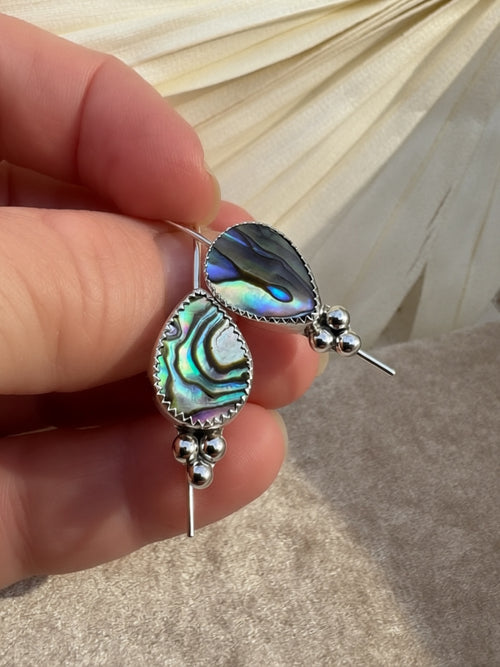 A close-up of a hand holding a pair of silver dangle earrings with teardrop-shaped abalone shell inlays that show iridescent green, blue, and purple hues, set against a neutral draped fabric background.