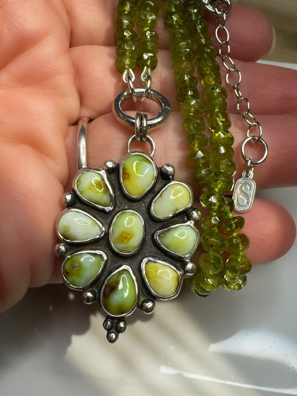 Close up of a hand holding the Verdant Sun statement necklace with nine green Palomino variscite stones, hand-knotted peridot beads and a sterling silver charm keeper.