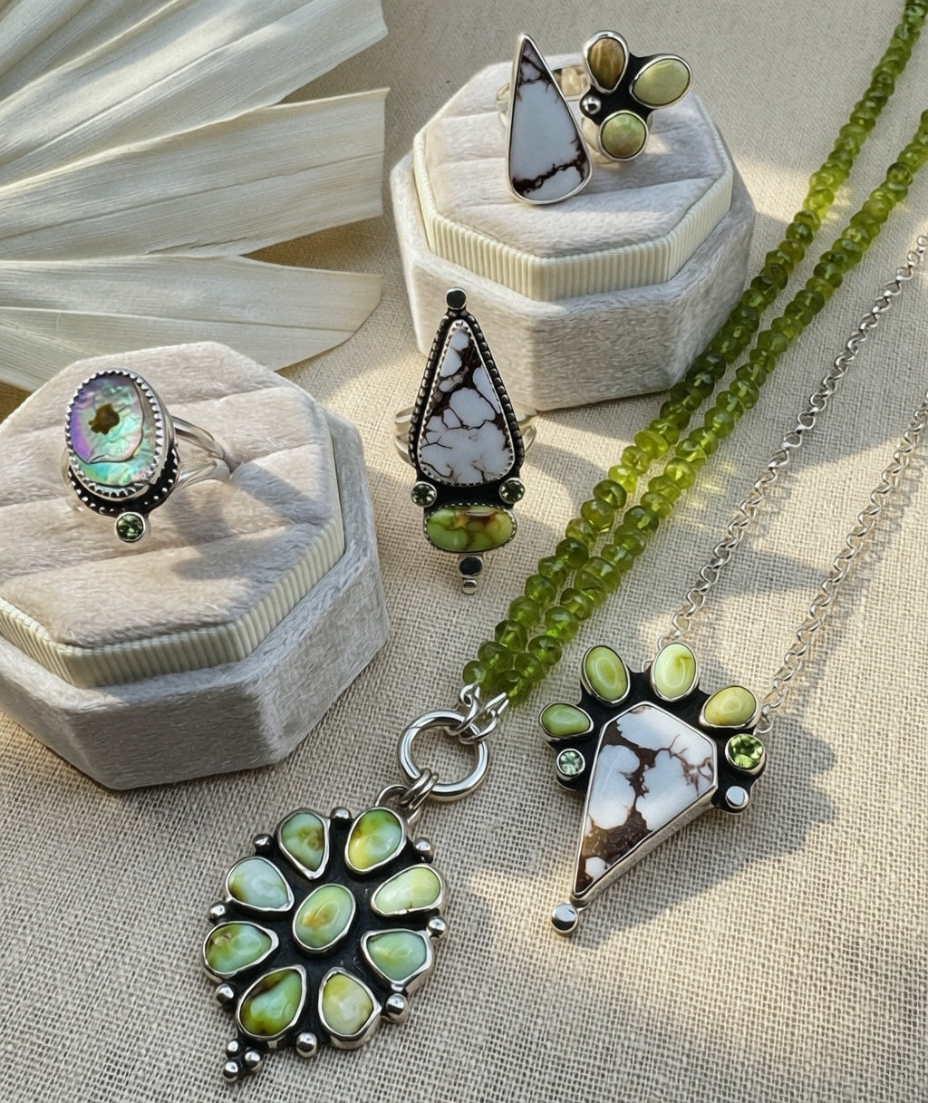 A styled jewelry flat-lay featuring multiple pieces of handmade jewelry, including a beaded green necklace, a silver ring with an iridescent stone, and a silver necklace with a large statement pendant, displayed on textured beige fabric and velvet jewelry boxes.