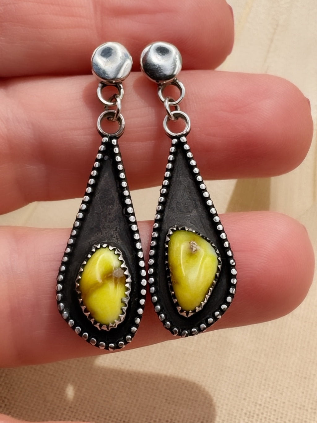 Close view of the earrings across fingers, showing the stones’ differing shapes, serrated bezels, and darkened silver recesses.
