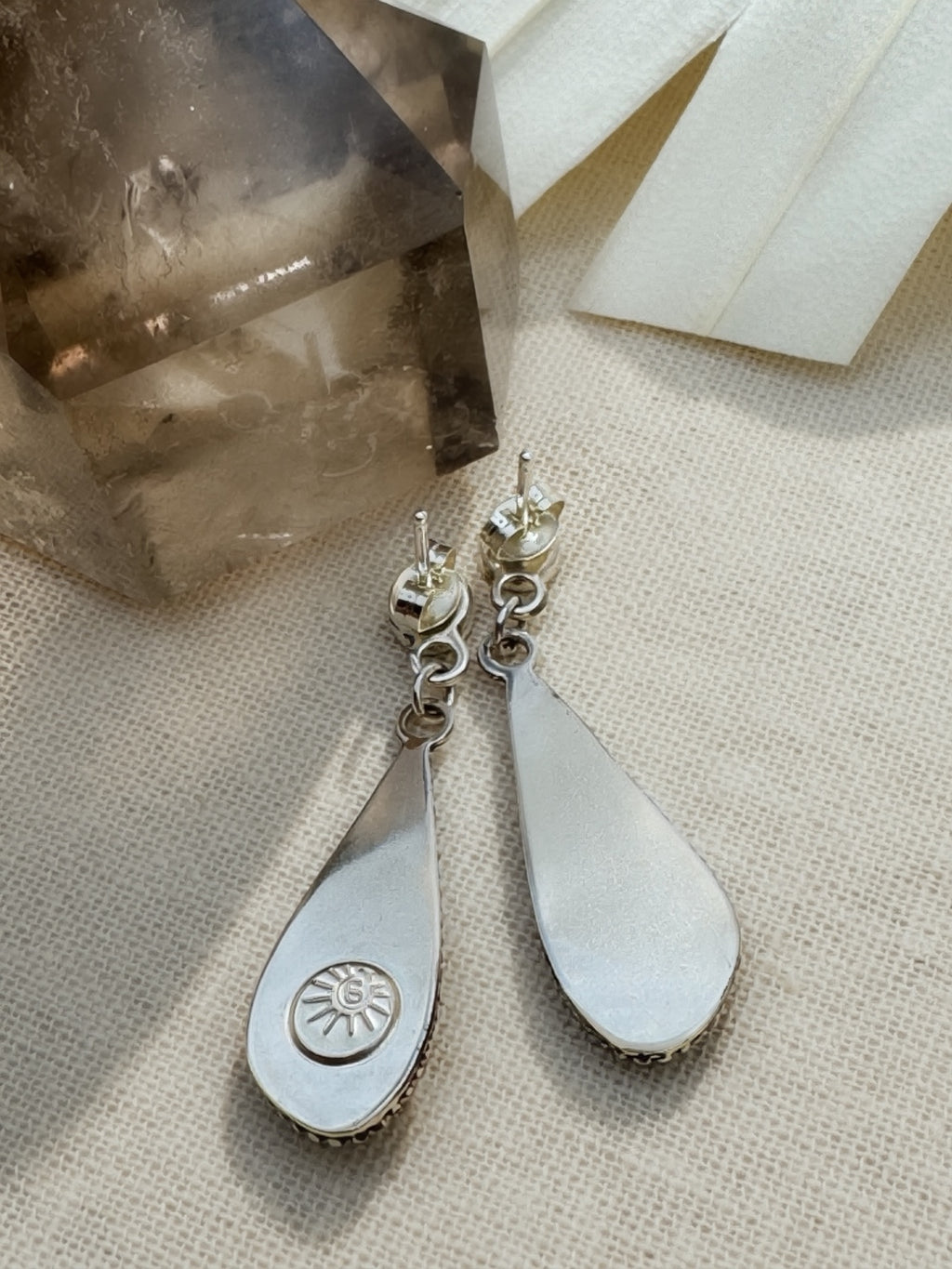 Reverse of the silver teardrop earrings on cream fabric, showing posts, ear nuts, connecting rings, and a sun-shaped maker’s mark on one back.