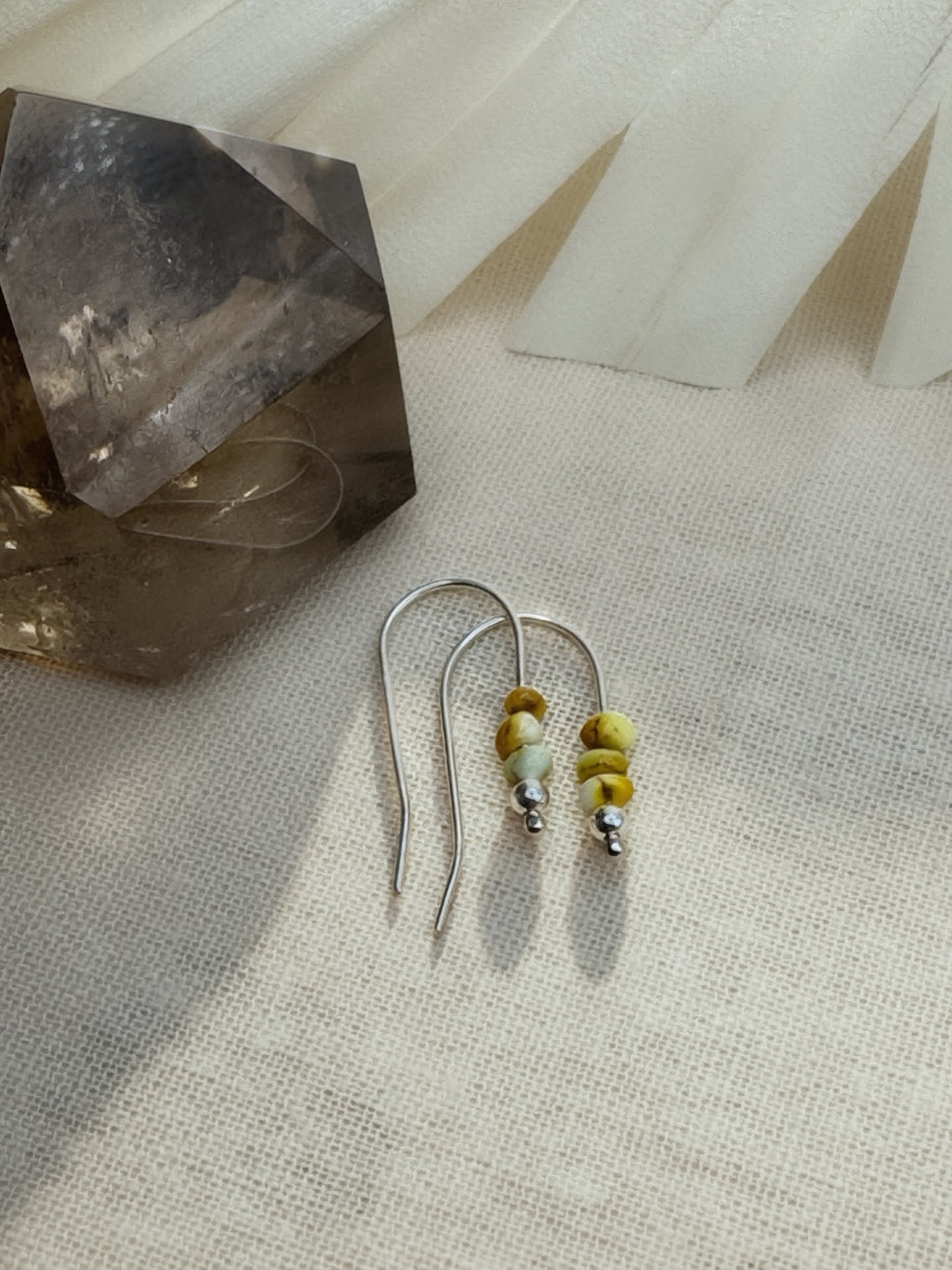 Full pair of Palomino variscite bead earrings on cream fabric beside a brown crystal, with long silver wire tails visible.