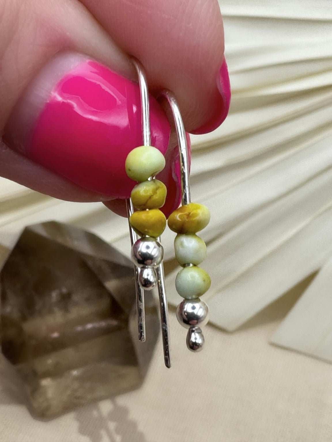 Close view of Palomino variscite earrings held between fingers, showing pale green and mustard-yellow beads above silver accents.