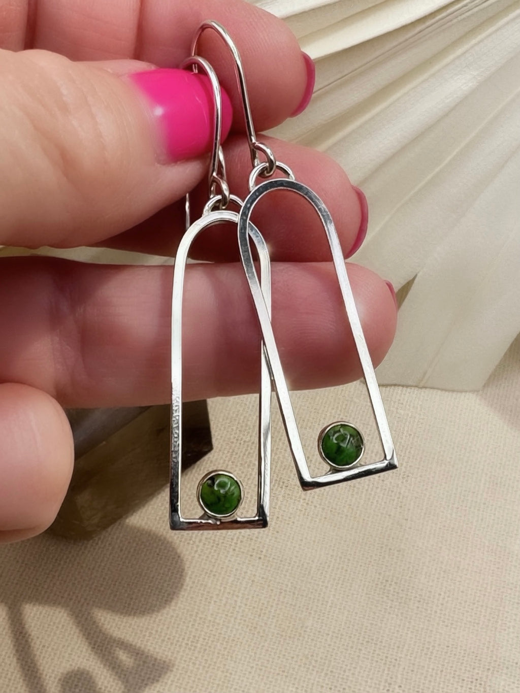 Sonoran Gold turquoise arch earrings held across fingers, showing green stone markings, slim silver frames, and curved ear wires.