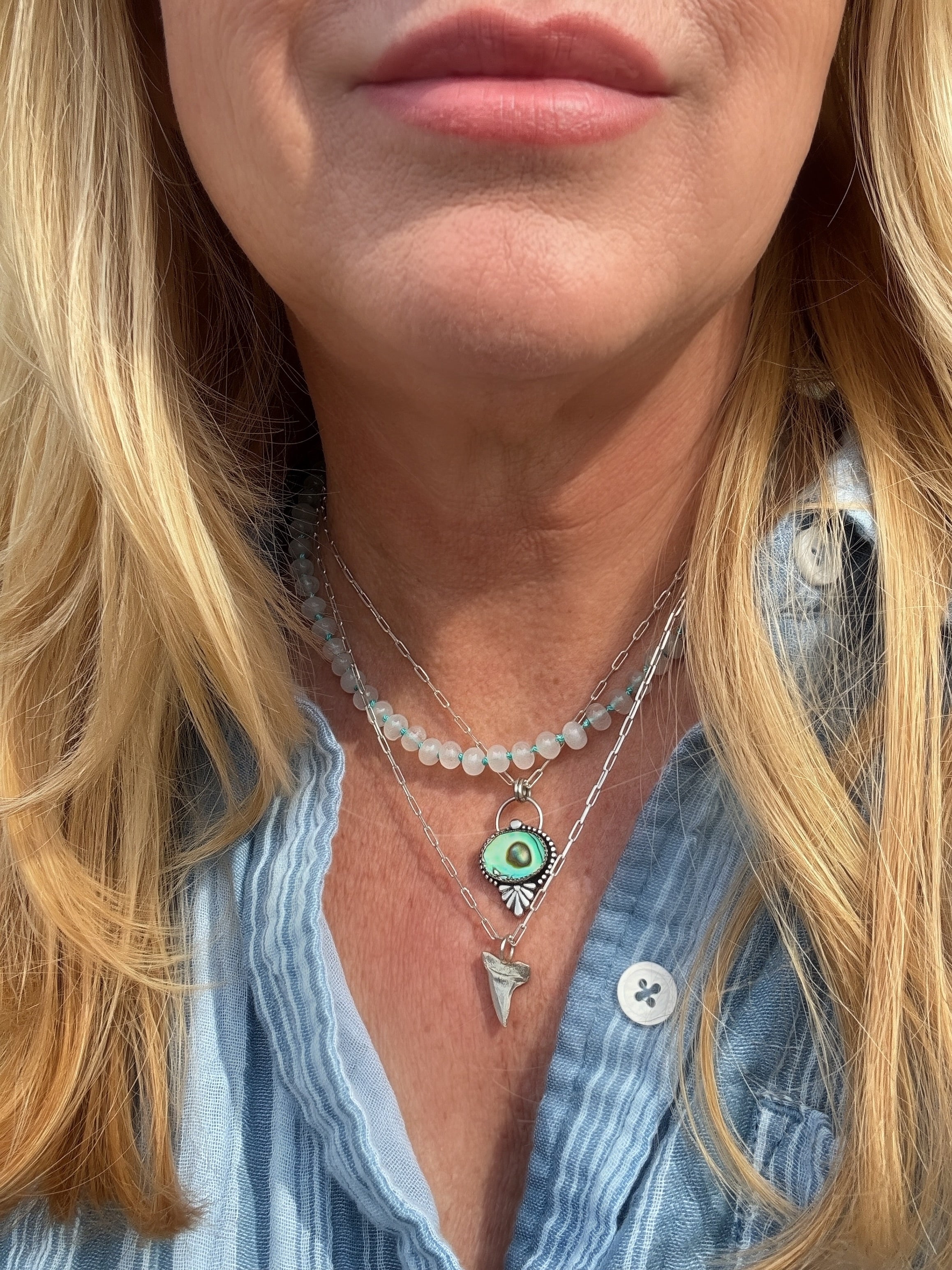 Close-up of candy jade rondelle beads hand knotted on turquoise silk thread stacked with an abalone shell necklace and a silver shark tooth necklace