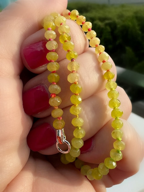Green beaded bracelet with rainbow knots, held in a hand with red nail polish against a blurred natural background