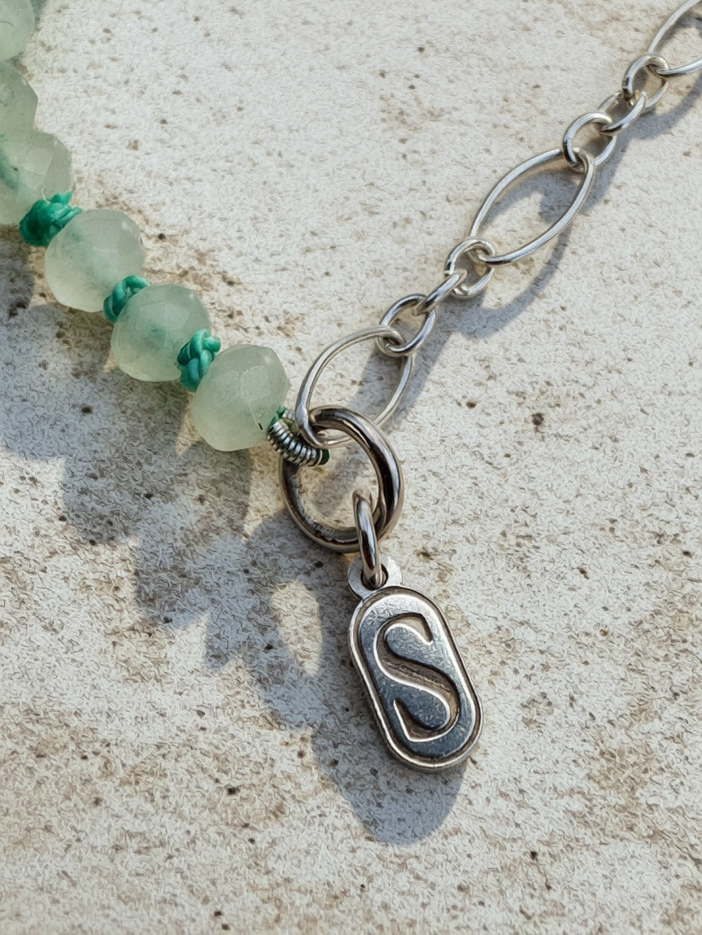 Close-up of a silver chain necklace with a polished oval pendant featuring a stylized letter 'S', attached to a strand of translucent pale green beads with small turquoise accents, laid on a light textured surface.