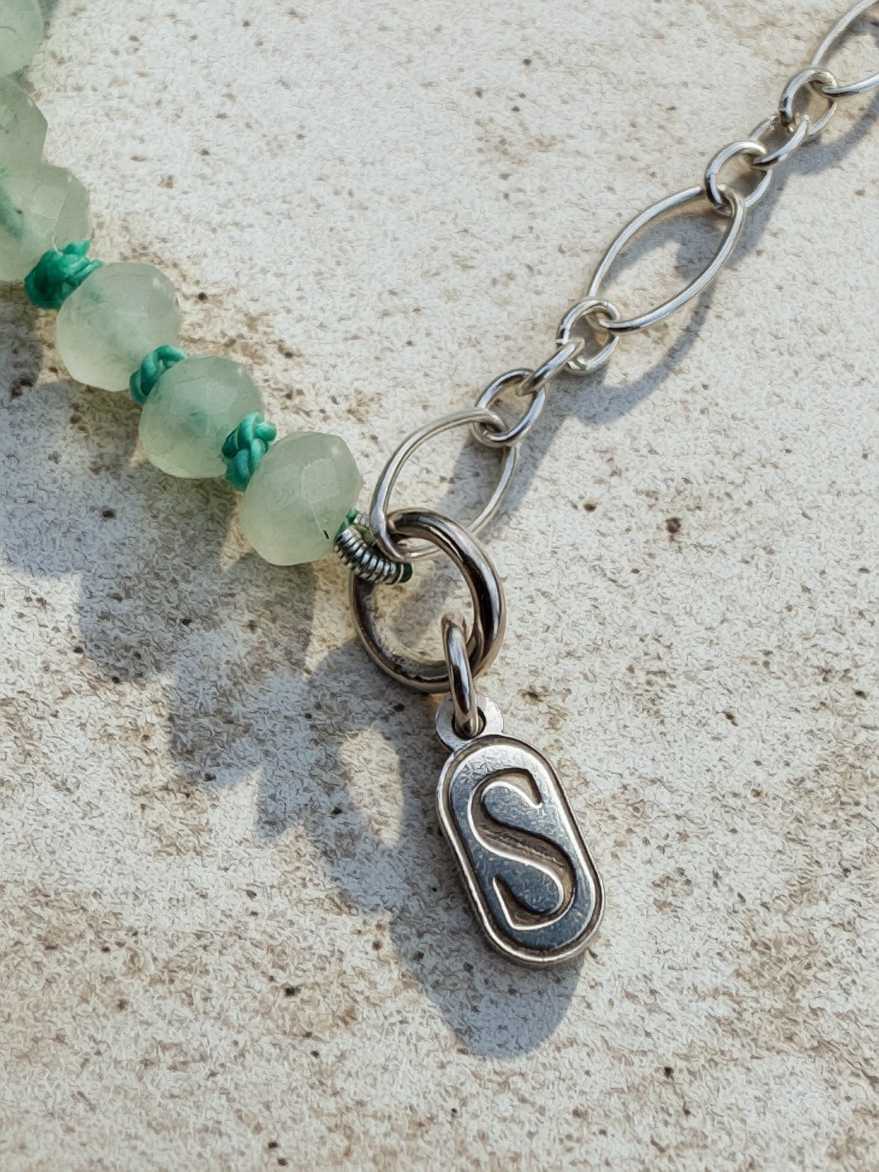 Close-up of a silver chain necklace with a polished oval pendant featuring a stylized letter 'S', attached to a strand of translucent pale green beads with small turquoise accents, laid on a light textured surface.