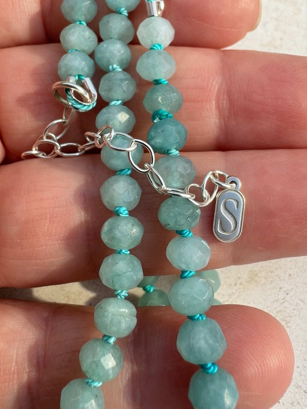 A close-up of a beaded necklace with faceted light aqua-green beads separated by bright turquoise thread, held in a person's hand. The necklace has a sterling silver-extender and a small oval silver charm engraved with an 'S'.