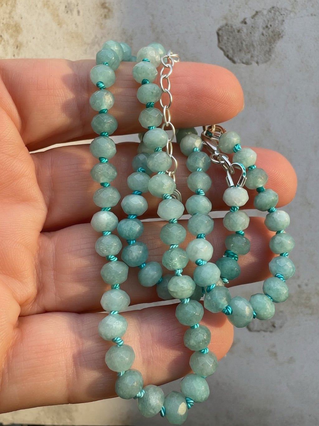 A close-up of a beaded necklace with faceted light aqua-green beads separated by bright turquoise thread, held in a person's hand. The necklace has a sterling silver-extender and a small oval silver charm engraved with an 'S'.