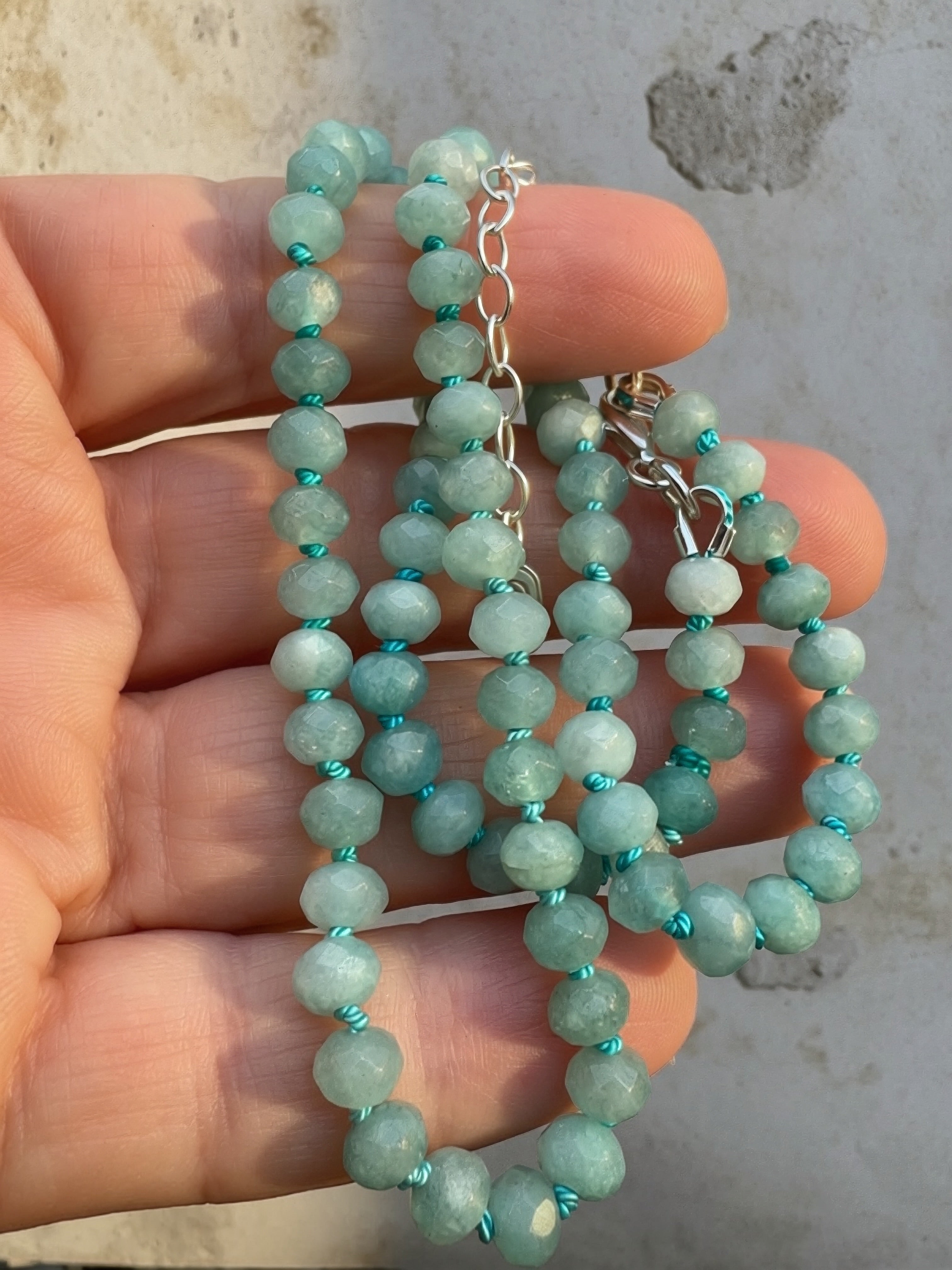 A close-up of a beaded necklace with faceted light aqua-green beads separated by bright turquoise thread, held in a person's hand. The necklace has a sterling silver-extender and a small oval silver charm engraved with an 'S'.