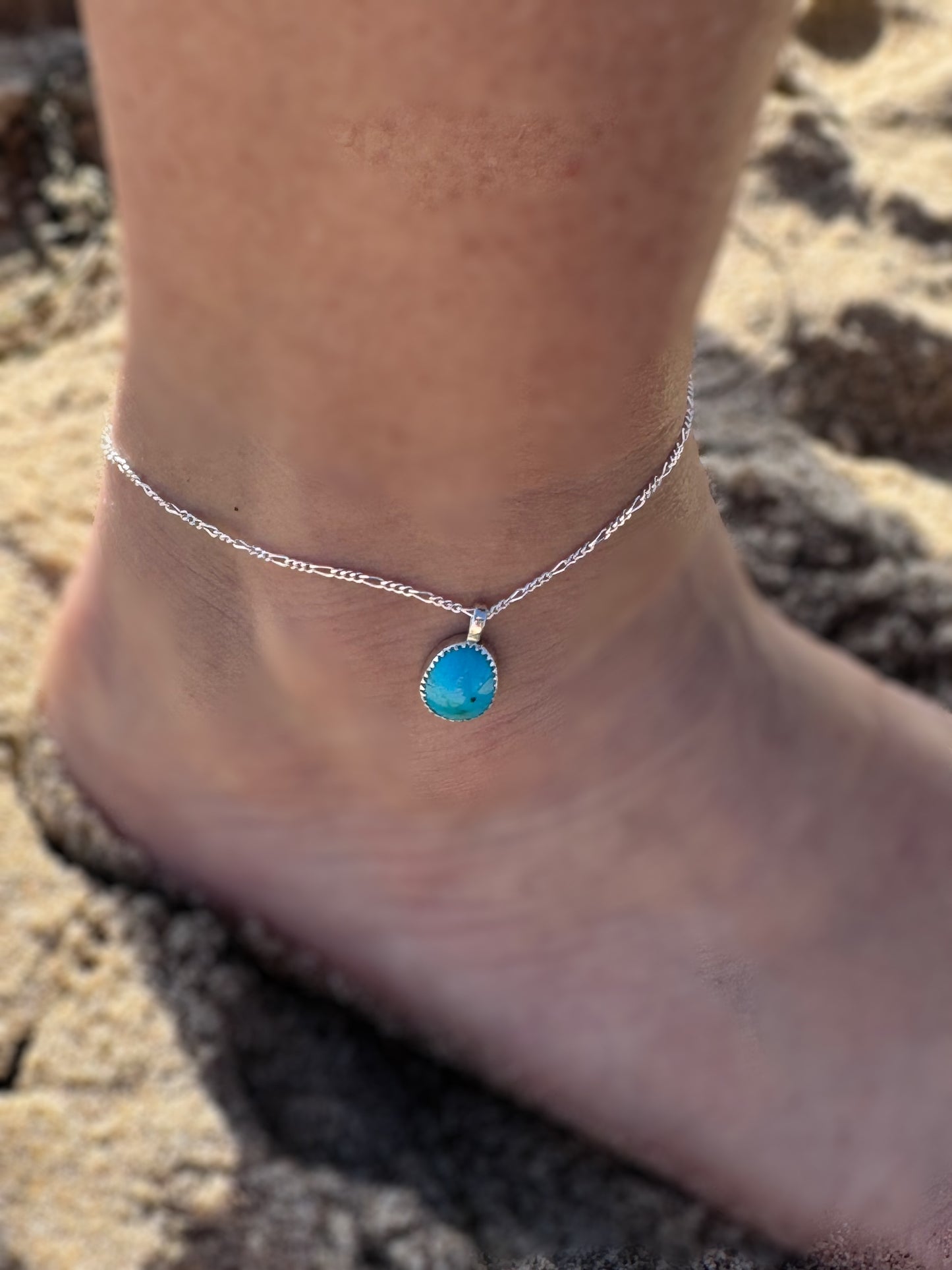 Tear drop shaped Bisbee turquoise anklet on a silver Figaro chain On an ankle against a sandy background