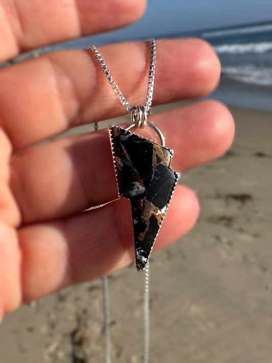 Hand holding a necklace with a unique lightning bolt - white buffalo and bronze - pendant against a beach background
