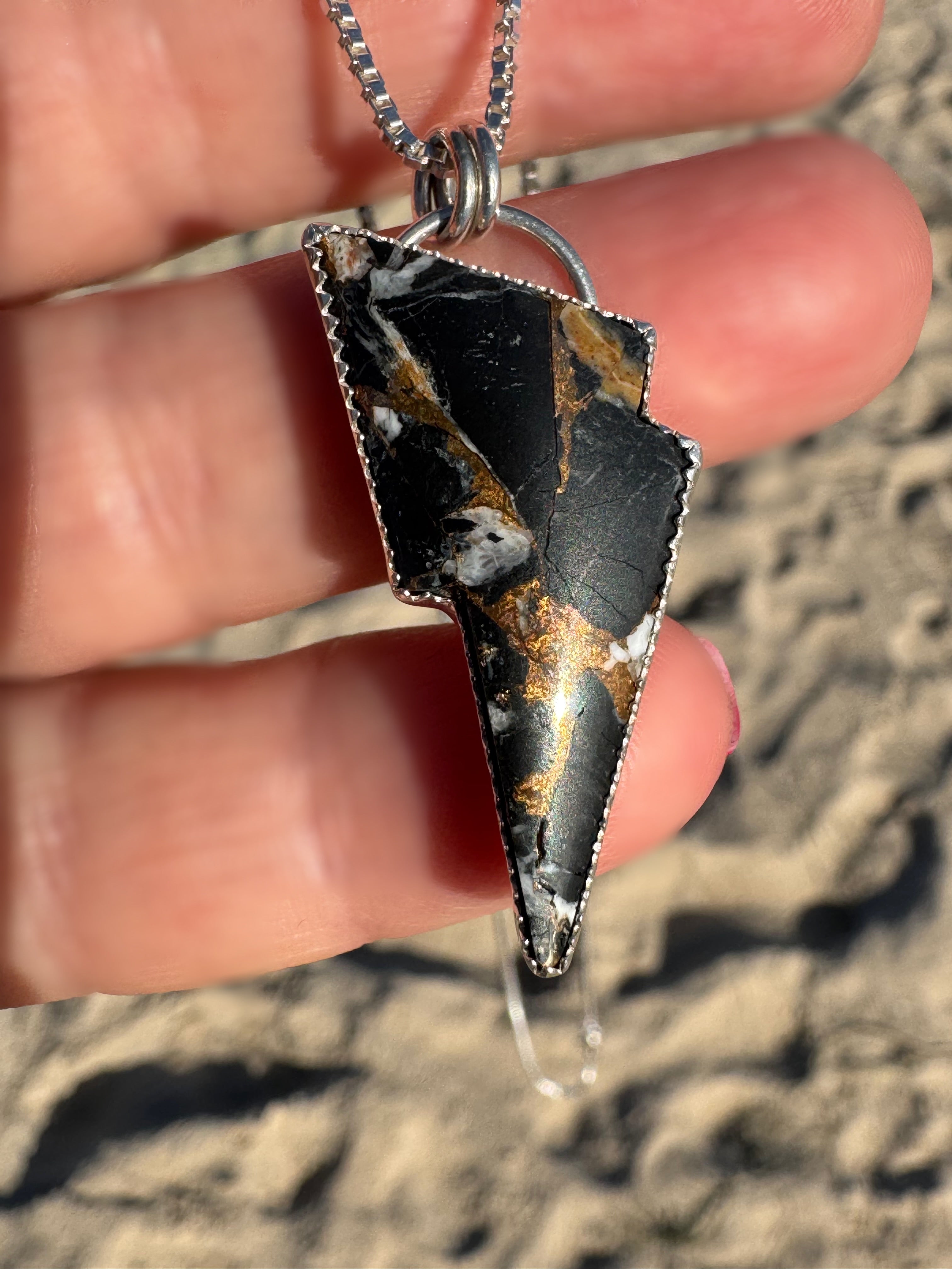 Pendant with a black and bronze/gold design held by a hand against a sandy background