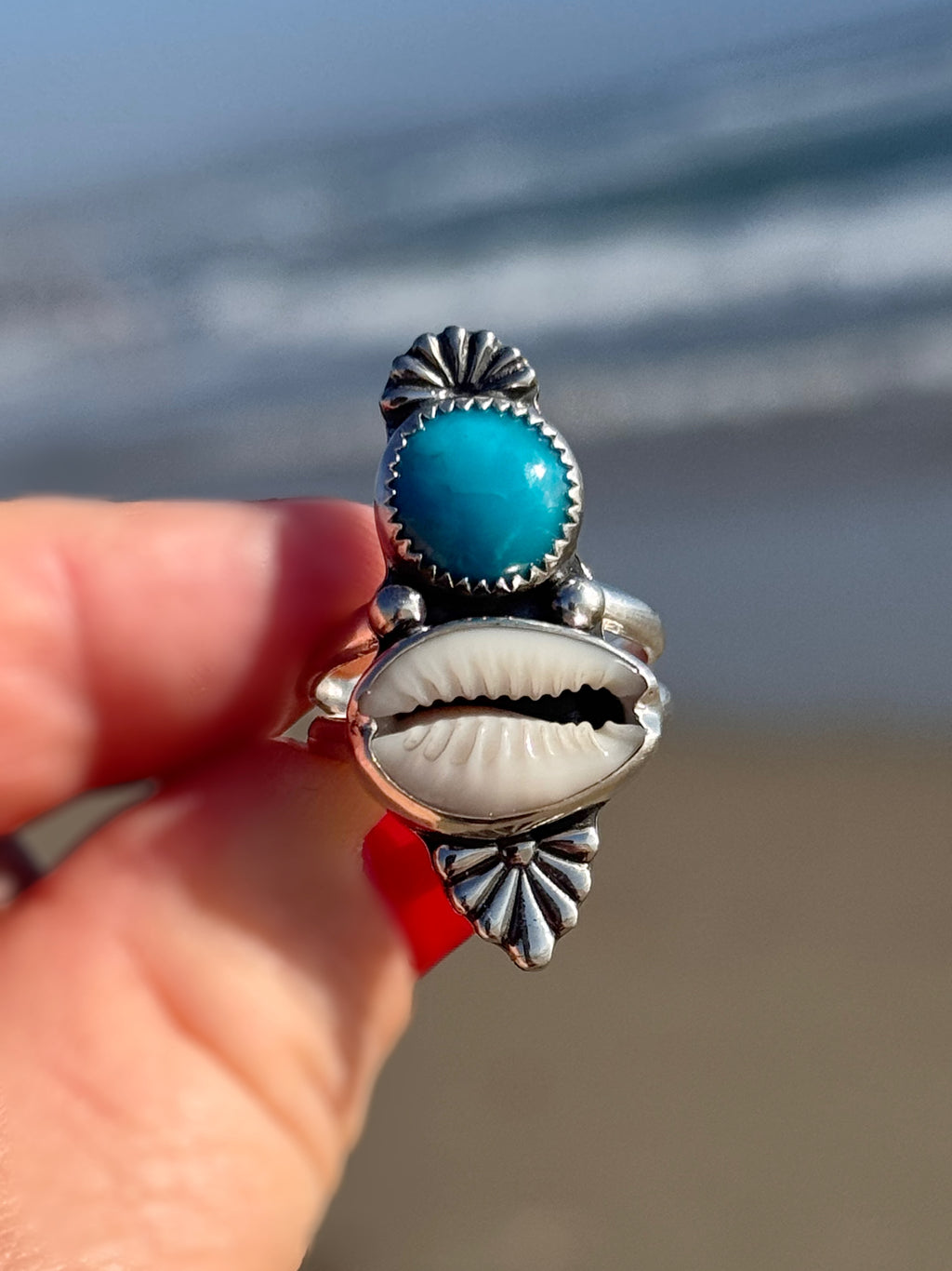 Silver ring with turquoise stone and cowrie shell held by a hand against a blurred beach background