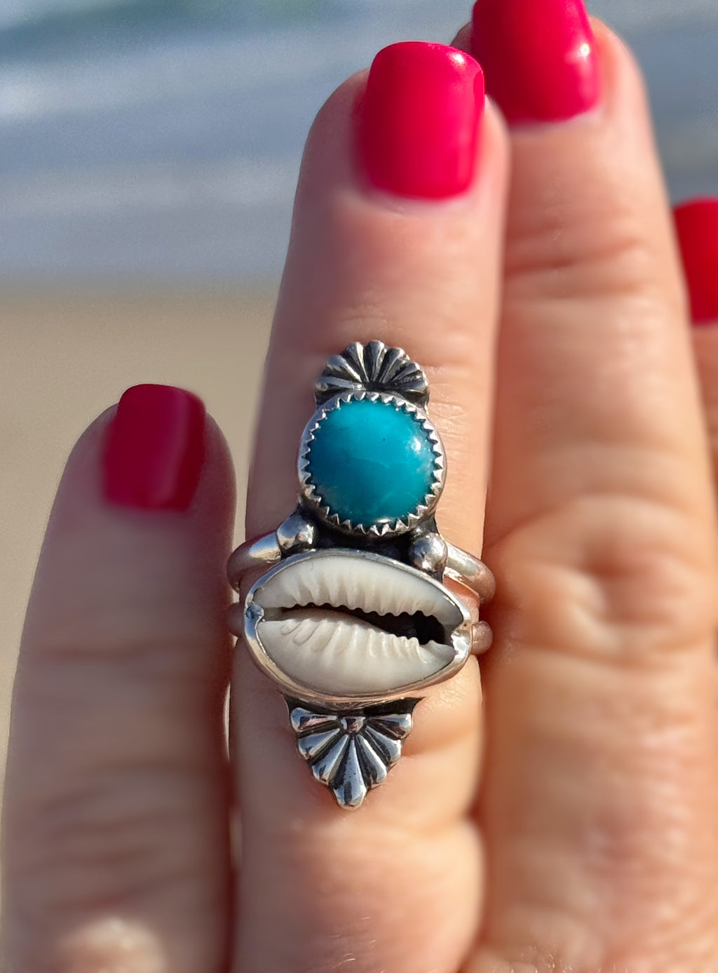 Silver ring with turquoise stone and shell design on a hand with red nails against a beach background