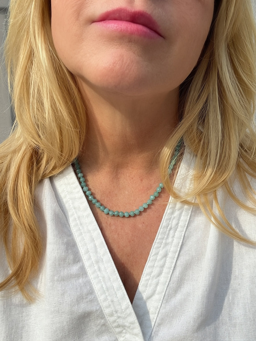 A close-up of a woman wearing a rainbow stone beaded necklace made of small round Caribbean-blue beads, paired with a white V-neck top.