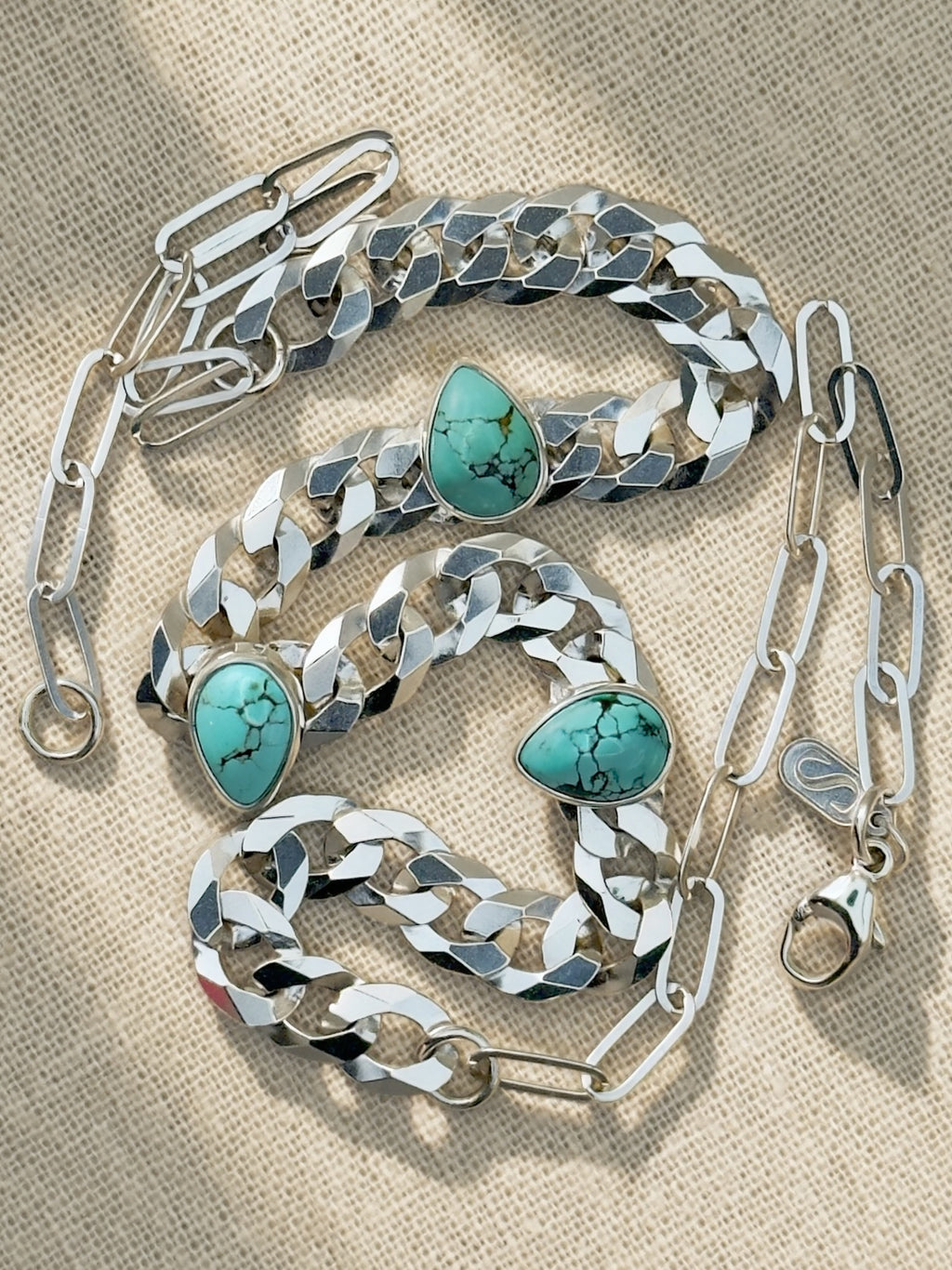 Full Blue Reign necklace coiled on cream fabric, showing curb and paperclip chains, lobster clasp, and maker’s tag.